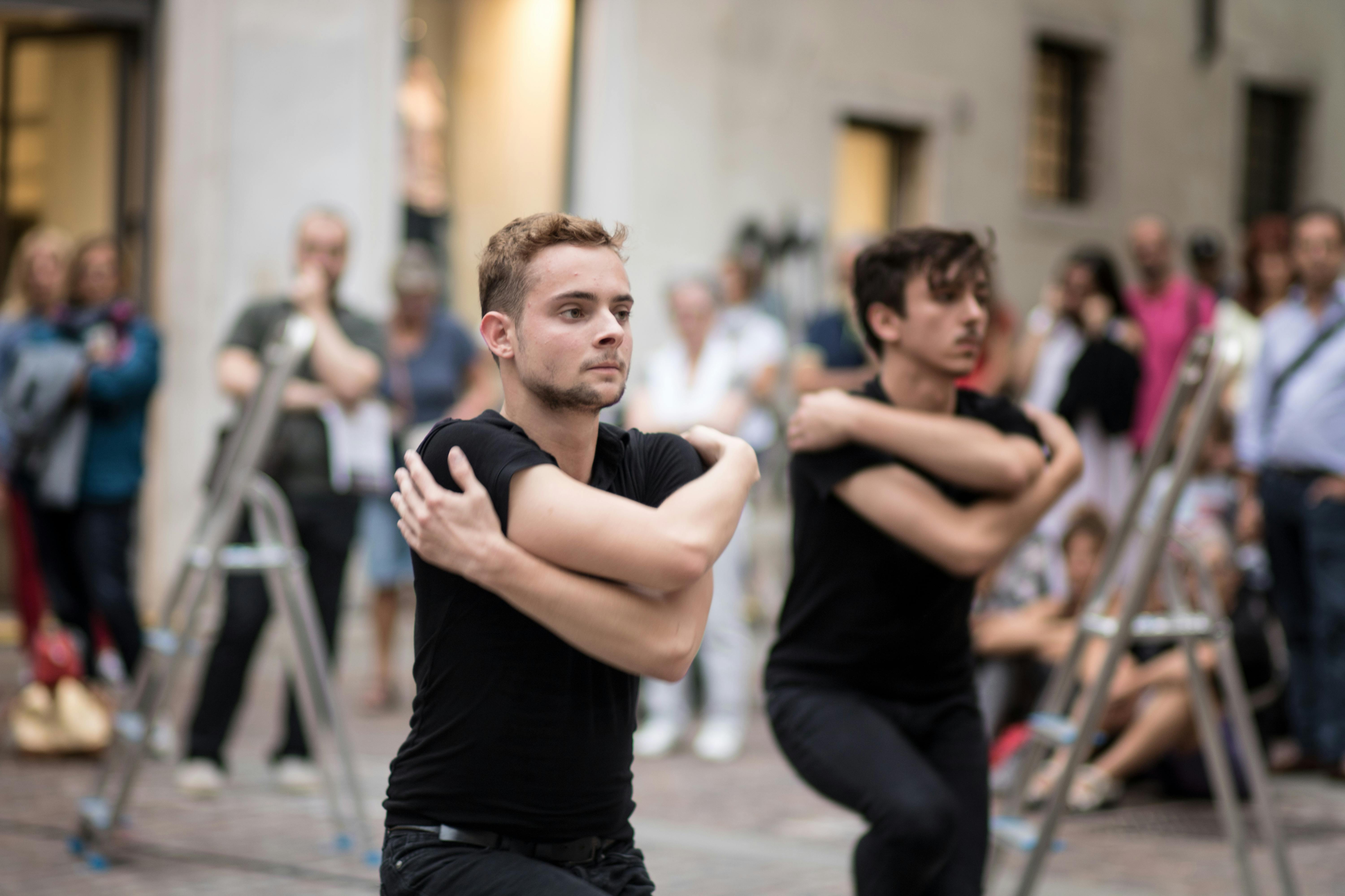 Due ballerini in primo piano si tengono le braccia intorno alle spalle con lo sguardo fisso in avanti