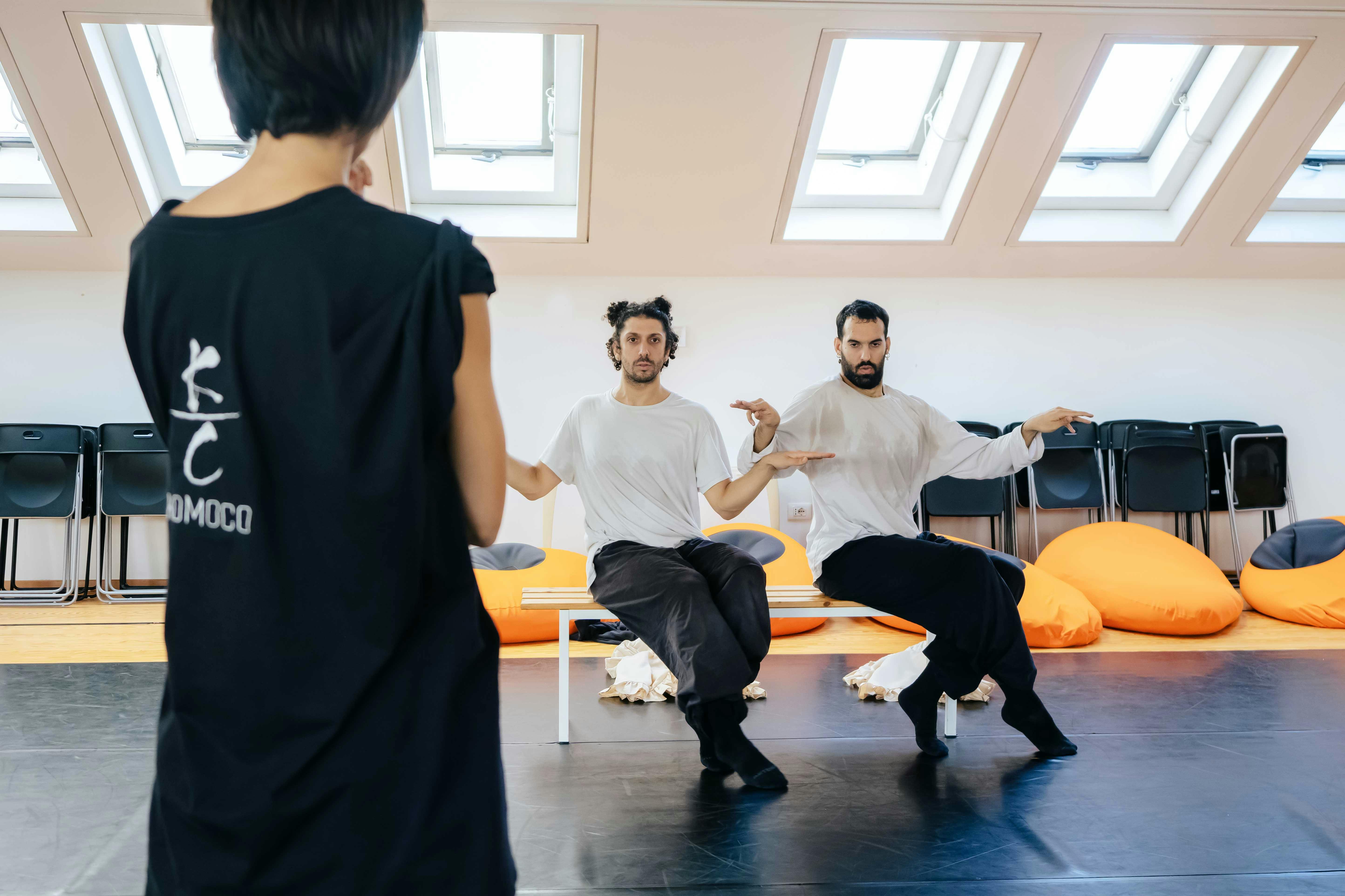 In primo piano Sofia Nappi di schiena con una t-shirt nera, in secondo piano i due danzatori in una posizione platica appoggiati a una panchina