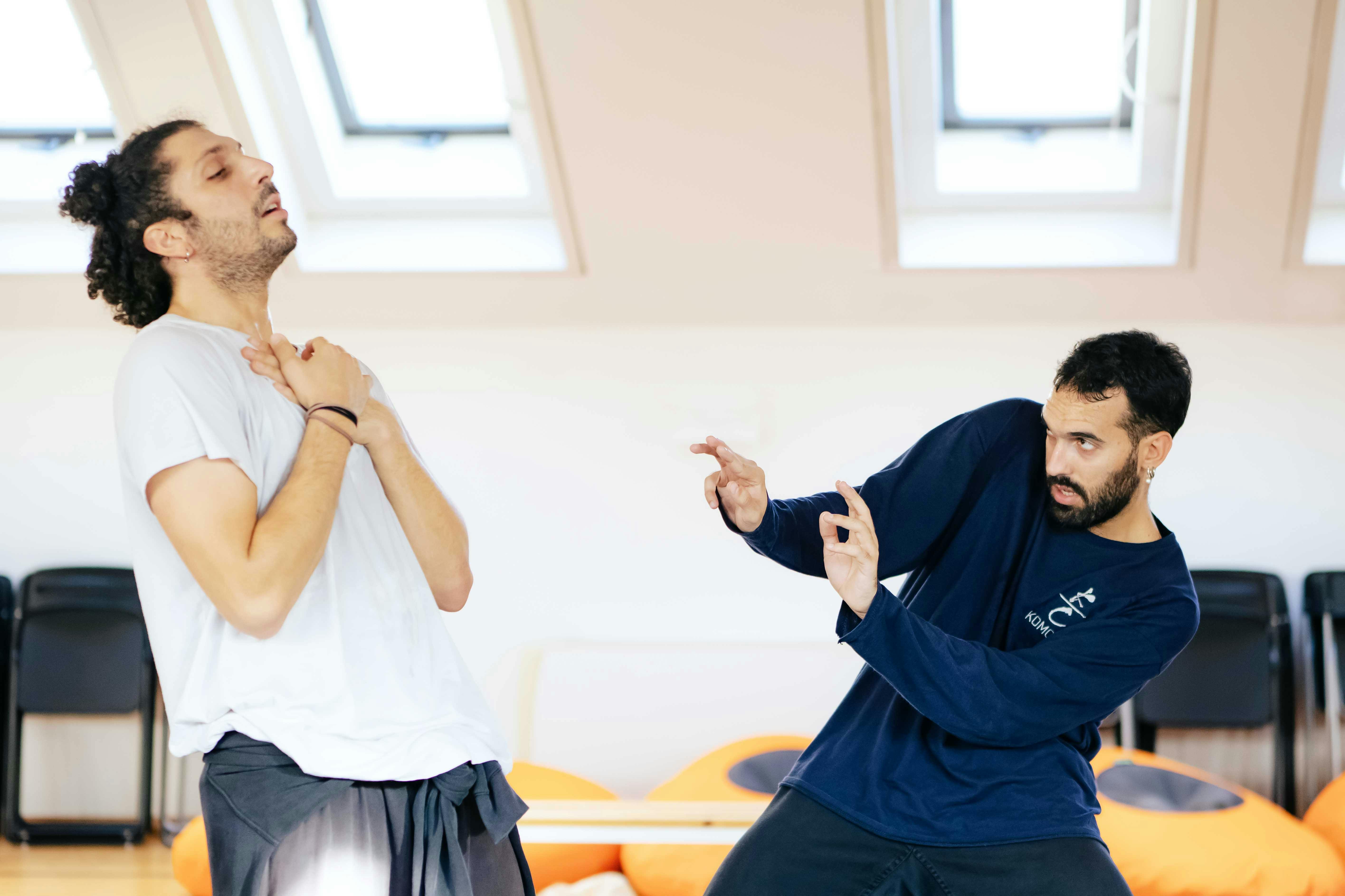 Un danzatore con t-shirt bianca tiene le mani al petto. Il secondo danzatore con maglia blu con schiena curva sula lato allunga le mani verso la prima figura.