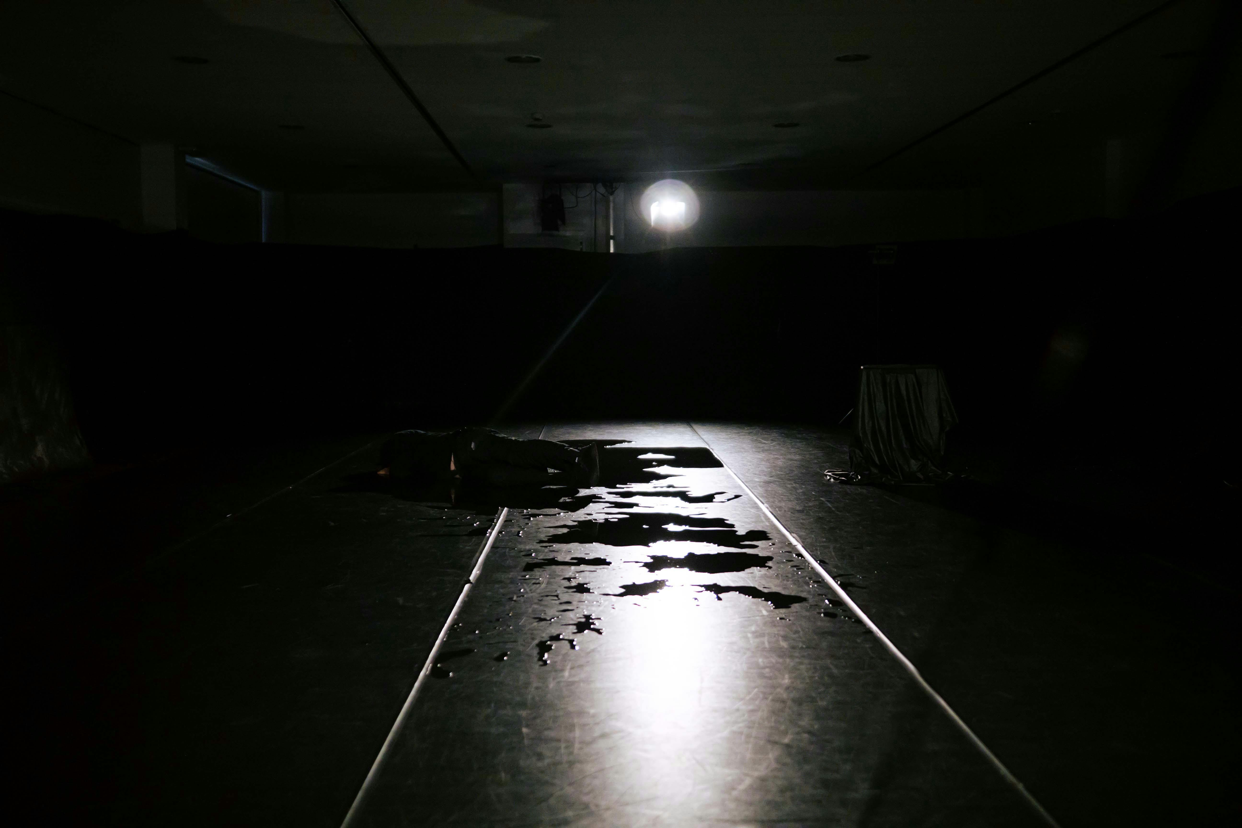Edoardo Mozzanega sdraiato sul fianco nella sala scura e vestito di nero, l'unica illuminazione è un faro in fondo alla sala che lascia delineare  la figura e delle macchie di acqua a terra