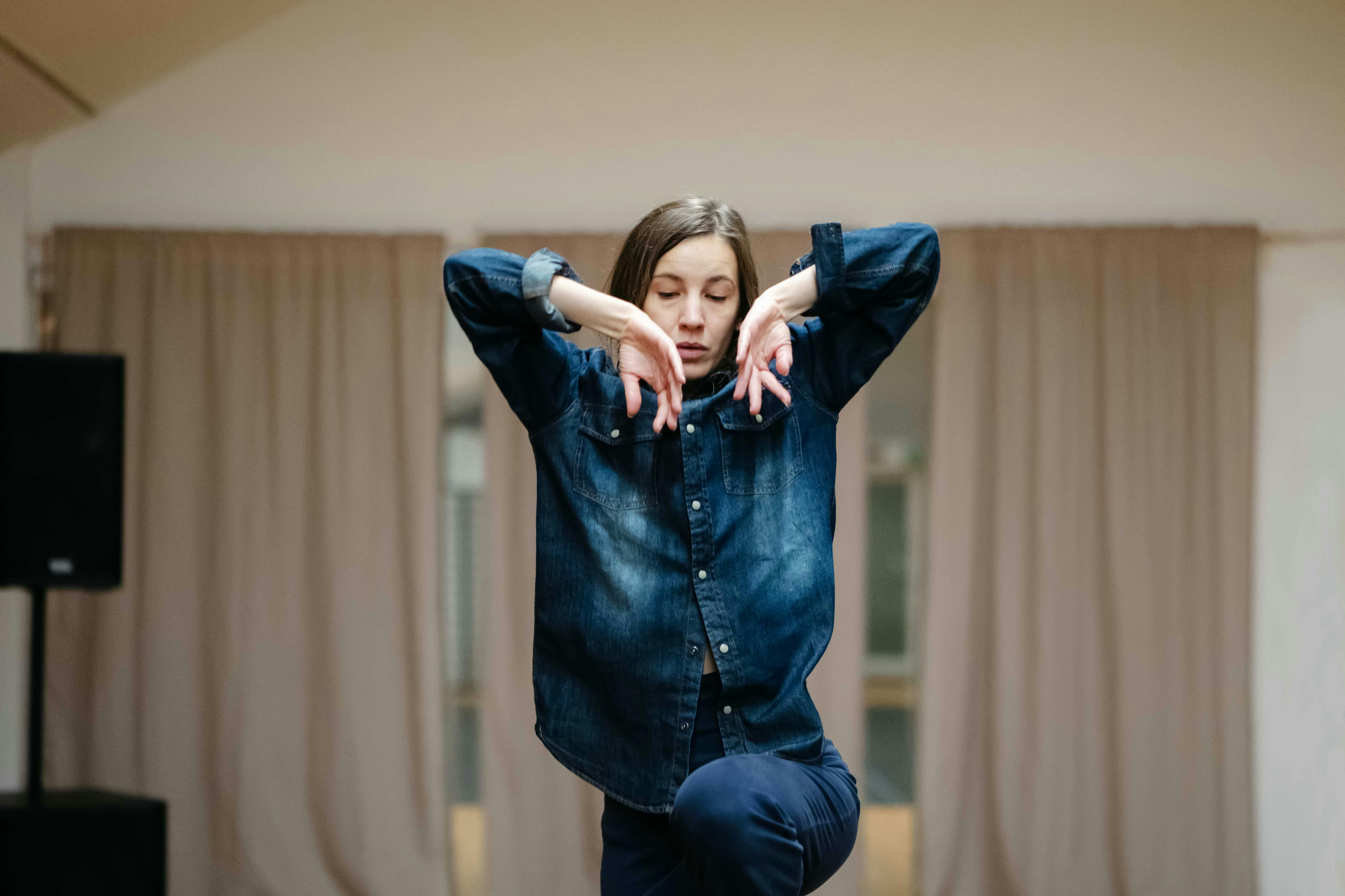 Stefania Tansini in sala danza indossa una camicia di jeans blu e pantaloni blu, le sue mani sono vicine al volto come per sfiorarlo con i gomiti alzati