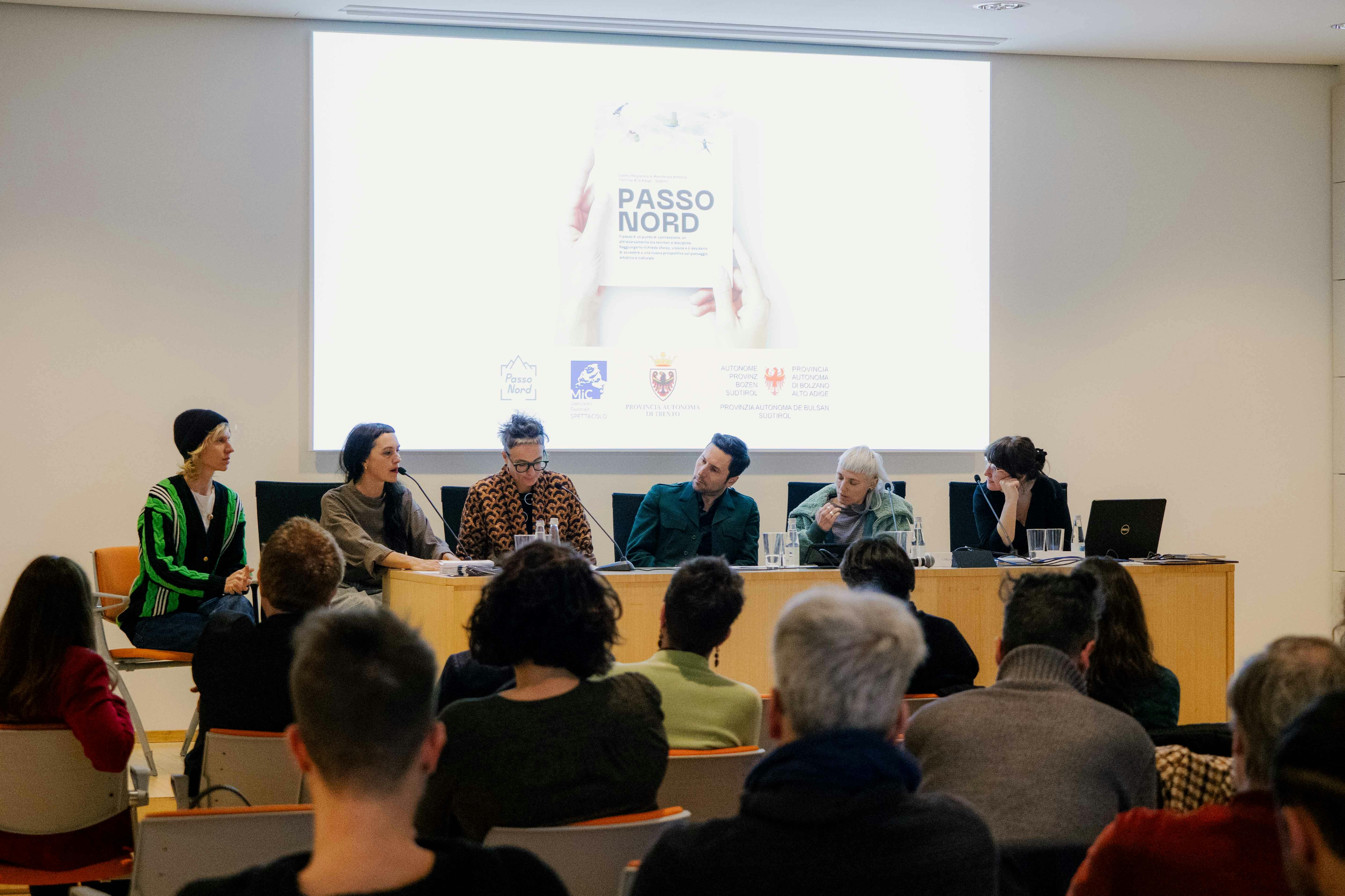 Sei persone tra artisti, artiste e operatori del settore seduti a un tavolo di una sala conferenze, di schiena il pubblico seduto ad ascoltare