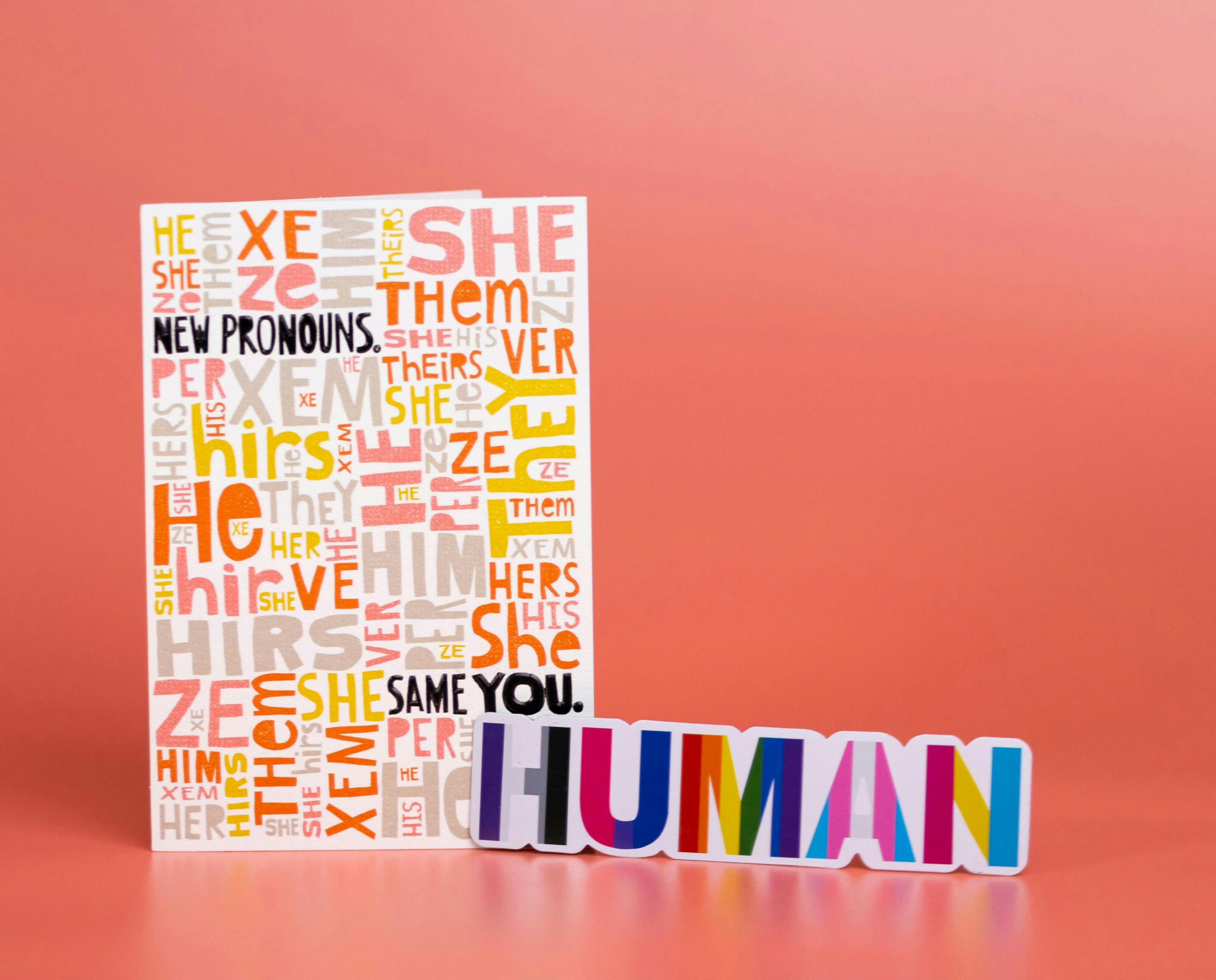 Su un foglio sono riportati in diverse lingue i pronomi che una persona può scegliere di usare. In primo piano, la scritta HUMAN ha i colori delle bandiere rainbow.