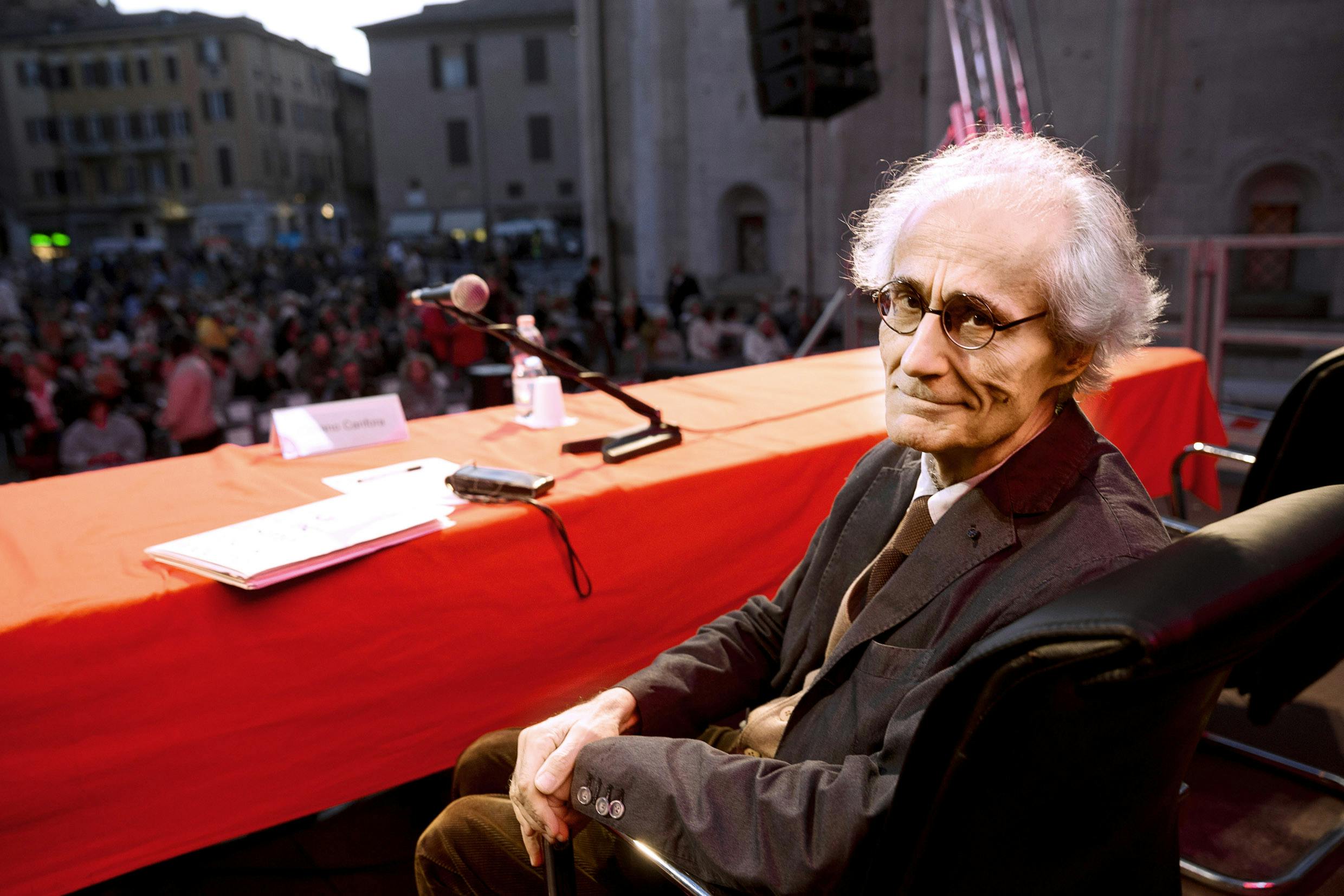 Luciano Canfora seduto ad un tavolo con un microfono davanti ad un pubblico.