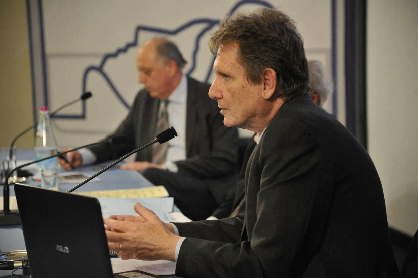 In the photo, Lucio Caracciolo, journalist. is on stage speaking into the microphone. He is wearing a dark suit.