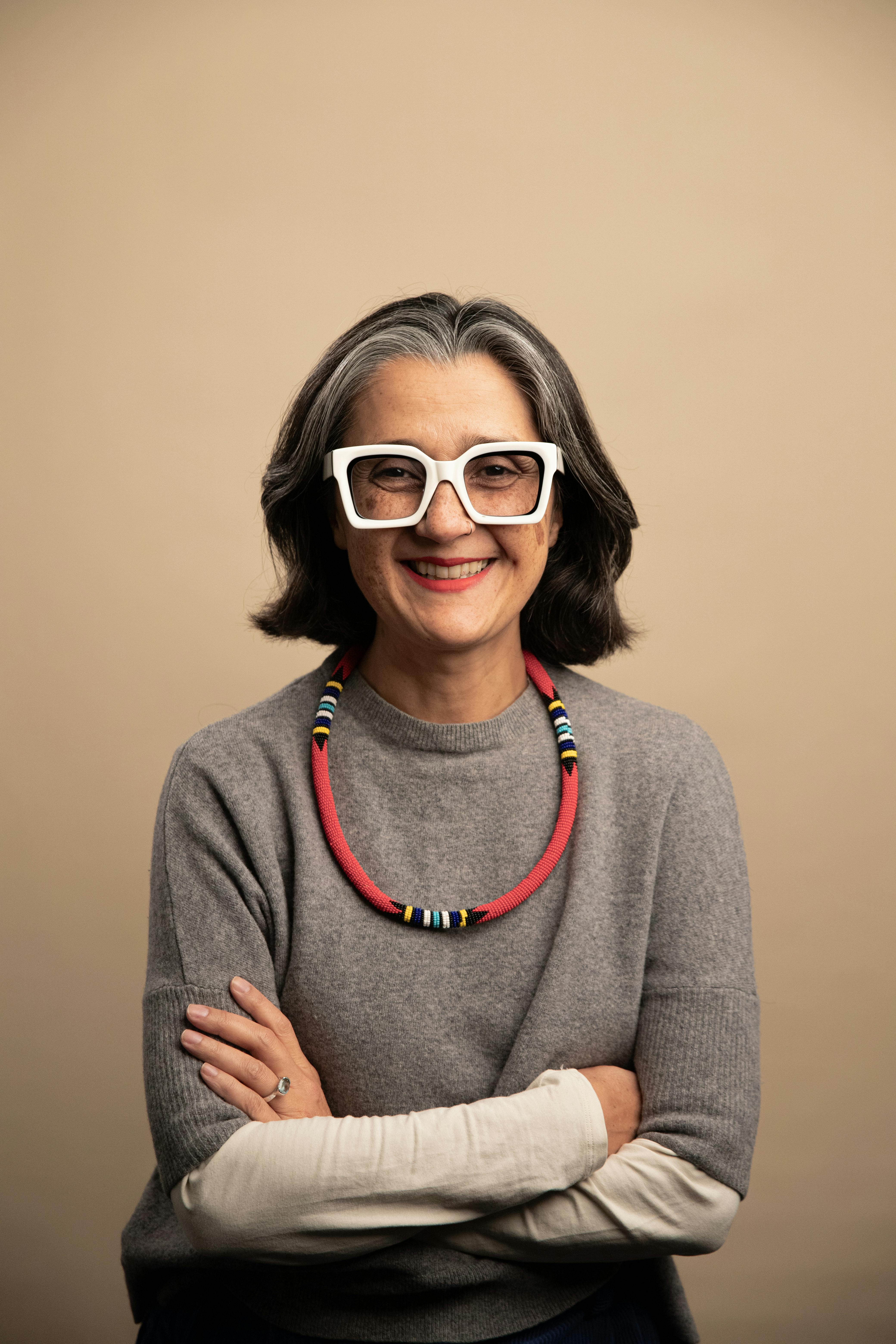 Portrait of Lorena Martínez Mier wearing flashy white glasses, a grey jumper and a colourful necklace