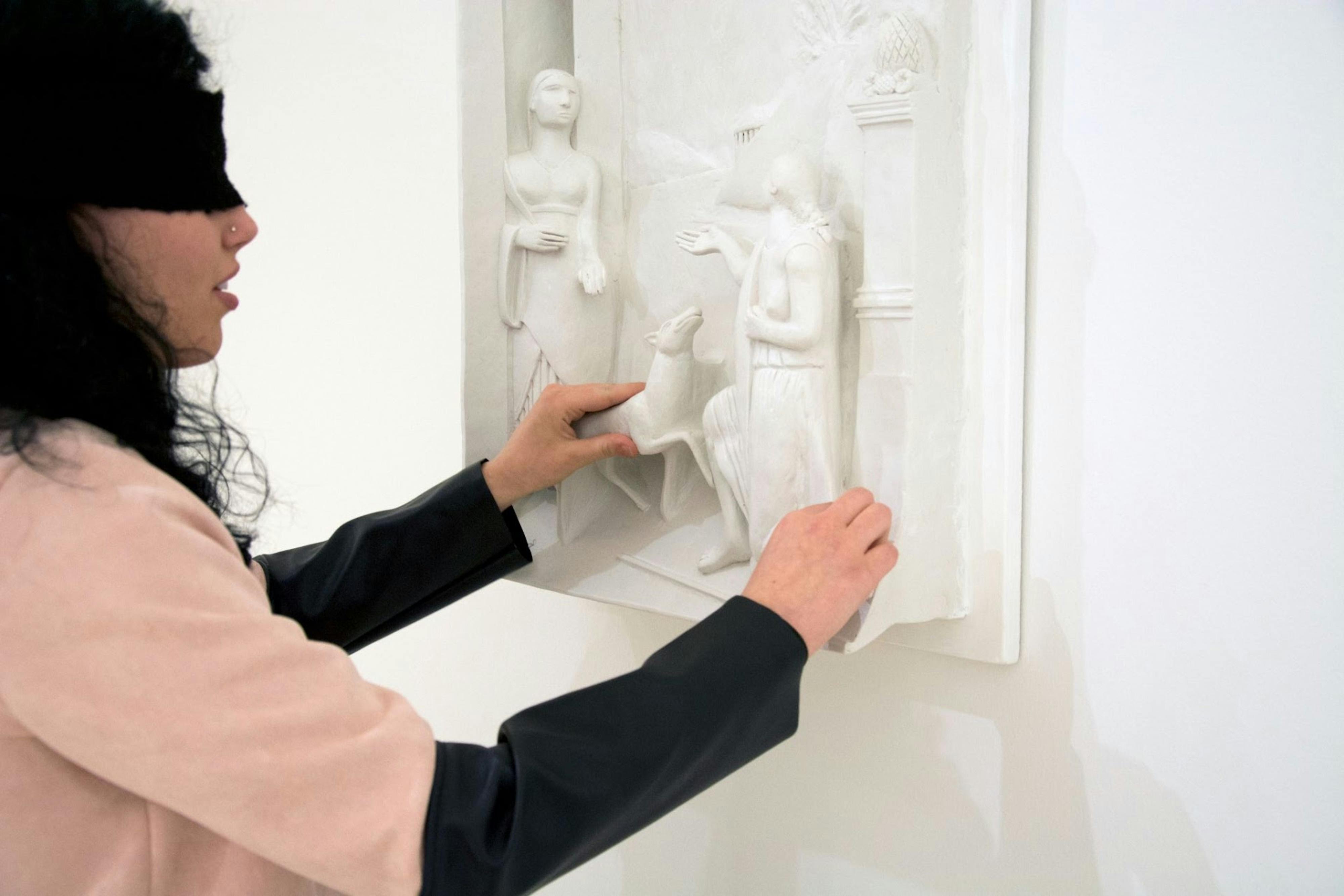 Una ragazza con una benda sugli occhi tocca l'altorilievo di un quadro