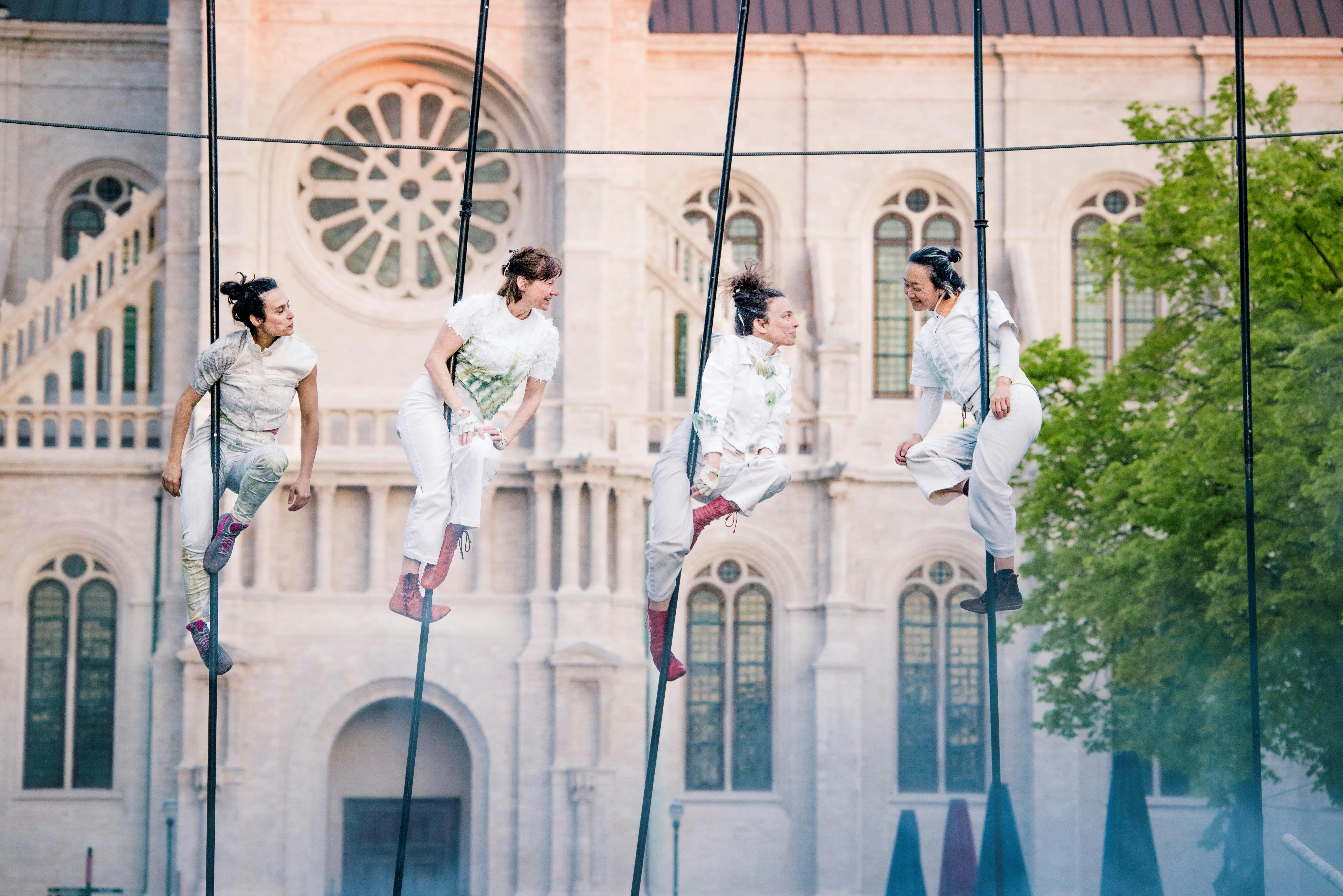 Four acrobatic performers dressed in white are suspended in the air, each holding onto a pole, and interact with each other.