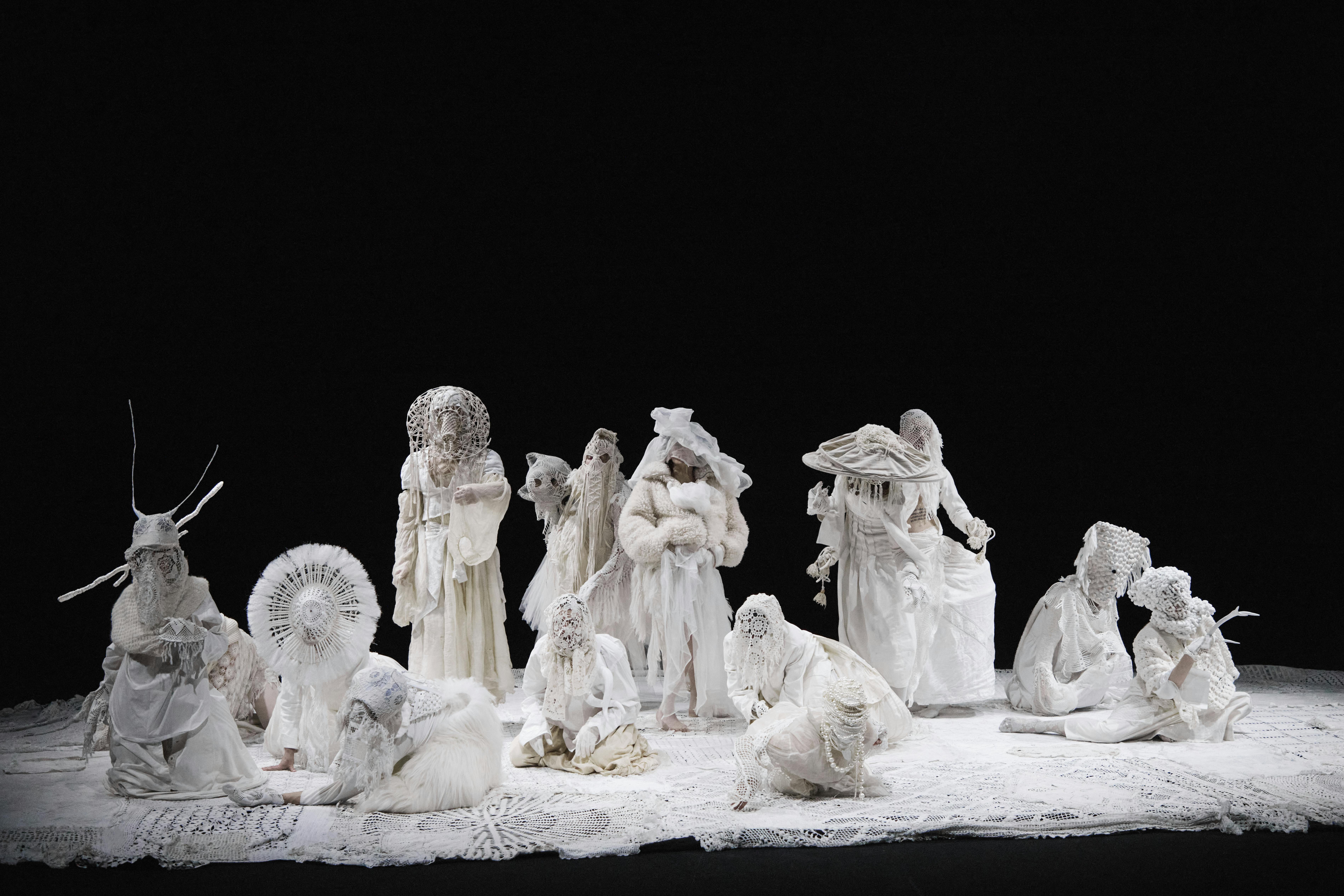 Ten performers on stage are dressed in white with lace costumes and frills that create animalistic and alien shapes. The stage is covered with the same fabric as the costumes, while the black background is in strong contrast