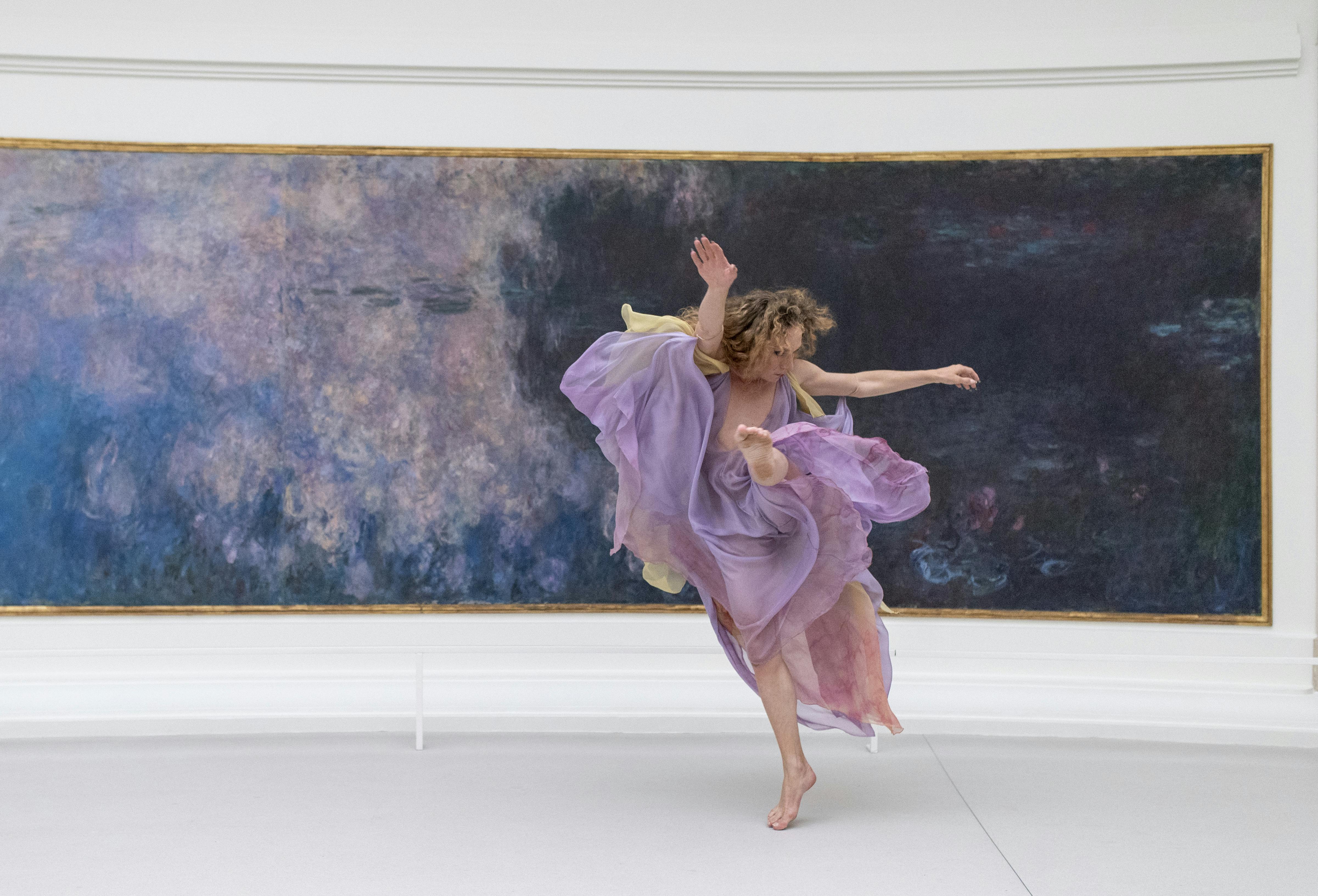 Performer dressed in light purple and yellow fabrics performs in a white museum room in front of a long horizontal painting. He has his arms raised and stands on tiptoe