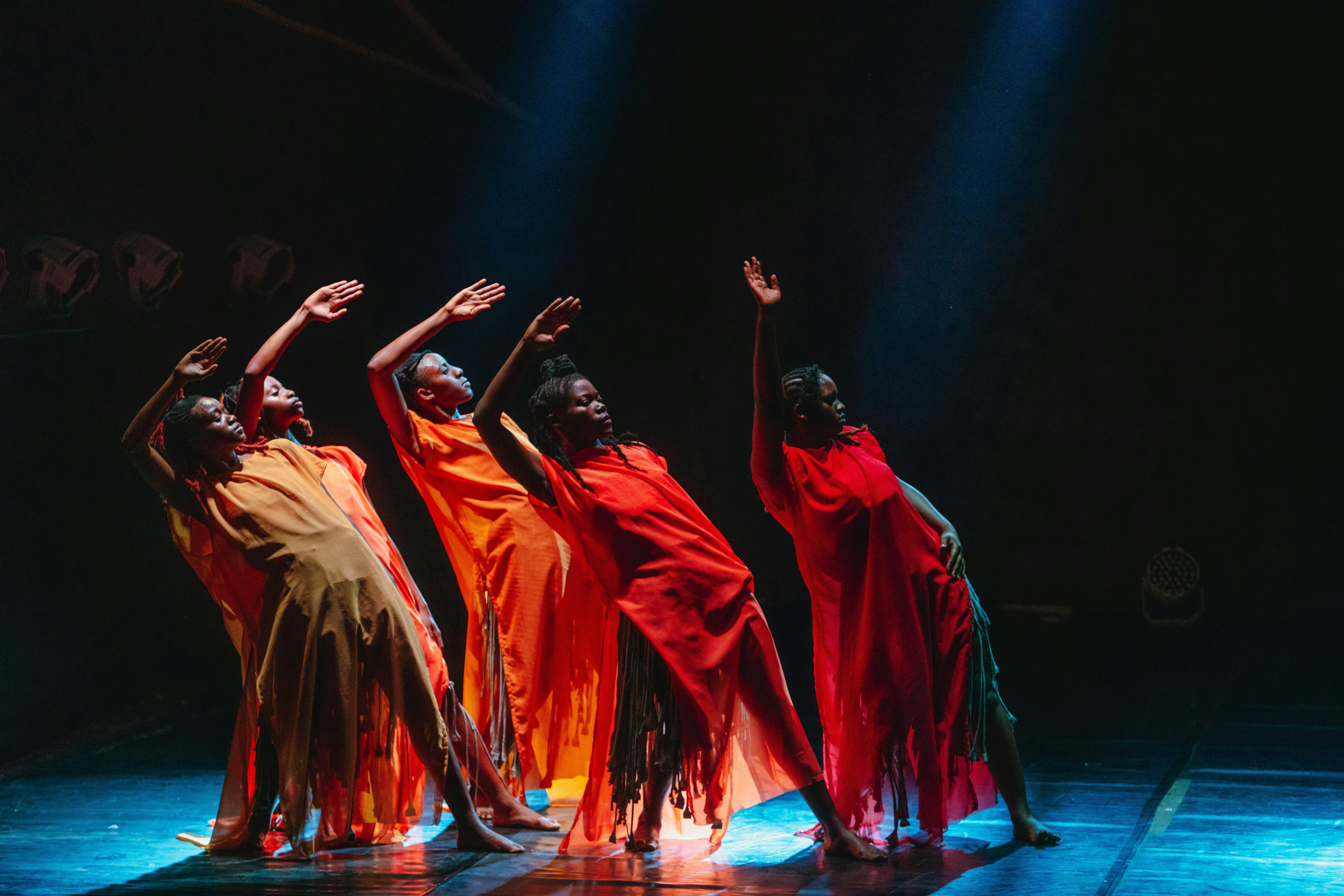 Cinque danzatrici indossano abiti sui toni del rosso e dell'arancione. Sono nella stessa posa, inclinate all'indietro, con una gamba tesa e un braccio alzato e piegato