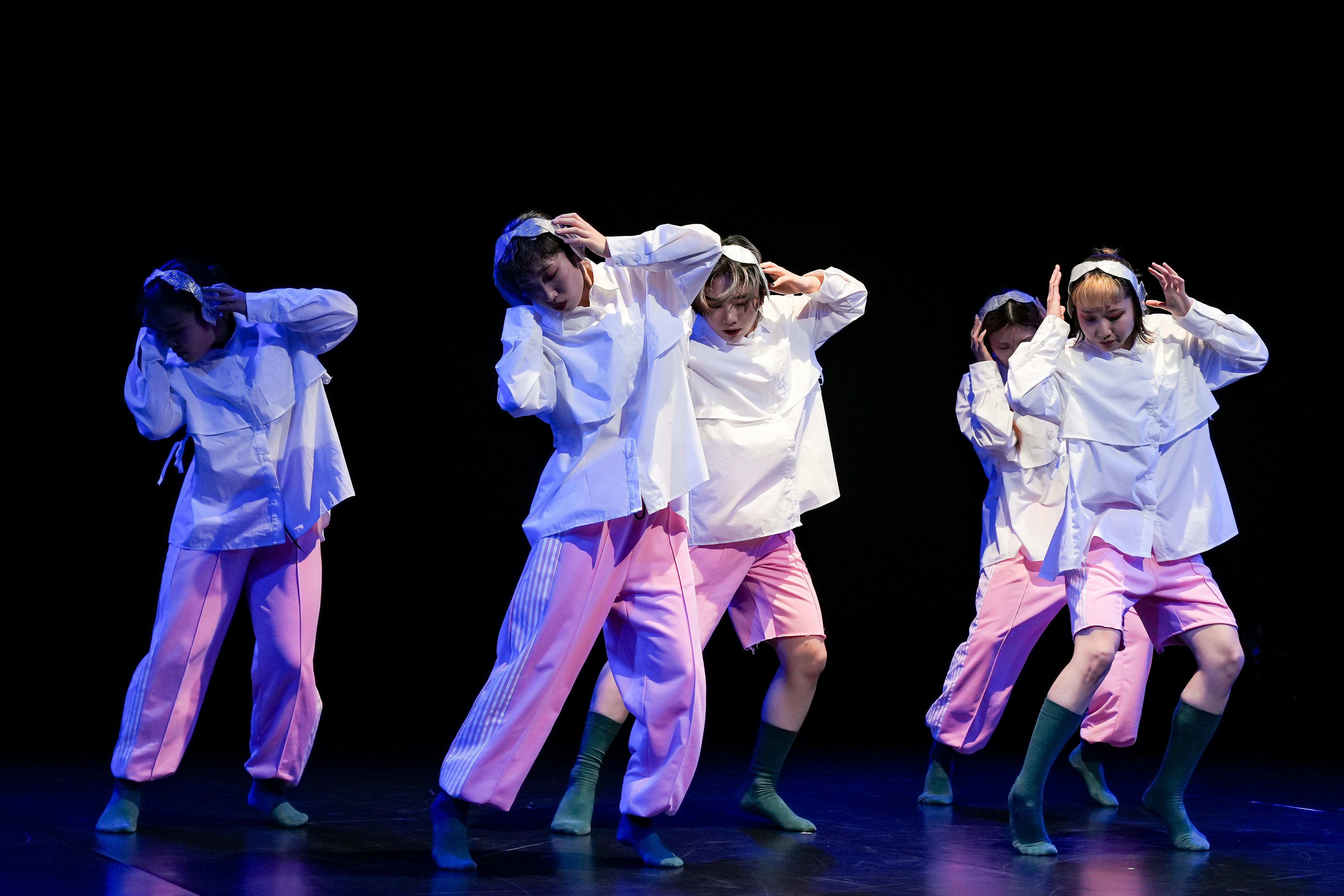 Five dancers on a dark stage are in the same pose with their faces turned downwards, their bodies slightly bent to the side and their hands to their ears. They are dressed the same with white shirts, pink trousers, green socks and earmuffs