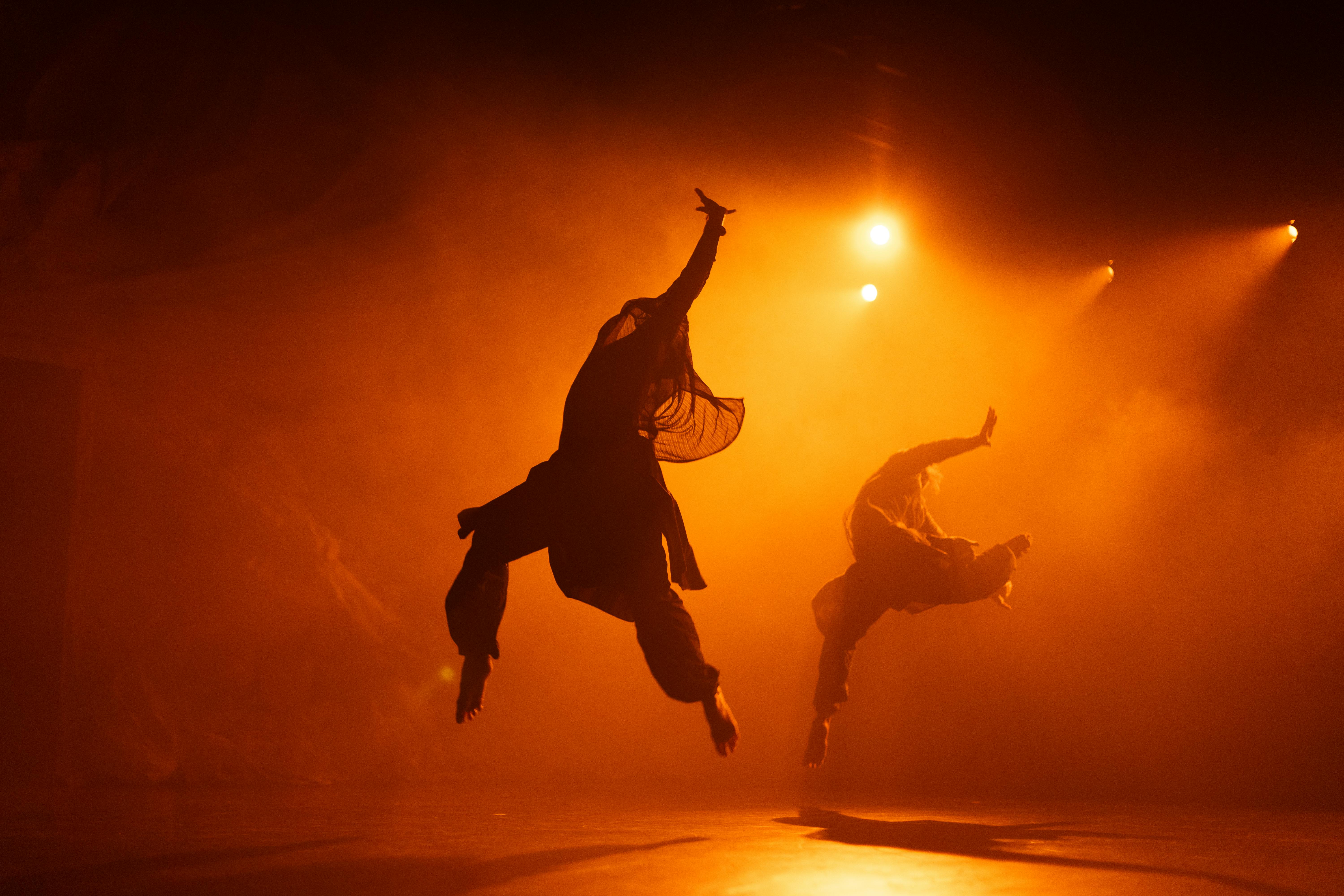 Two dancers in the air with one arm raised and legs apart. They wear loose and light clothes. Their bodies become silhouettes thanks to the orange light that hits them from behind