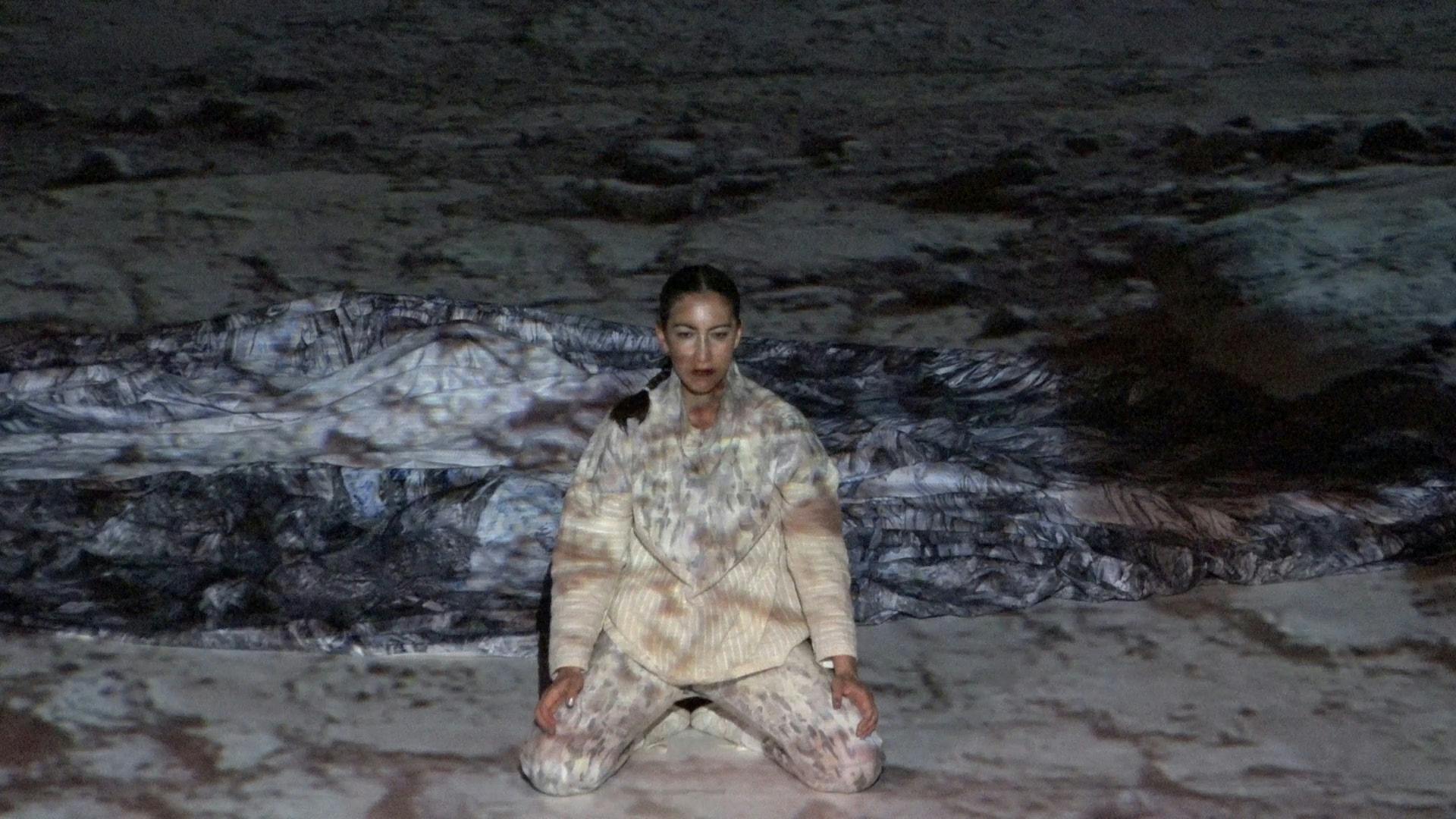 The artist Amanda Piña on stage wearing a white suit, a texture is projected, she is kneeling and looking at the audience