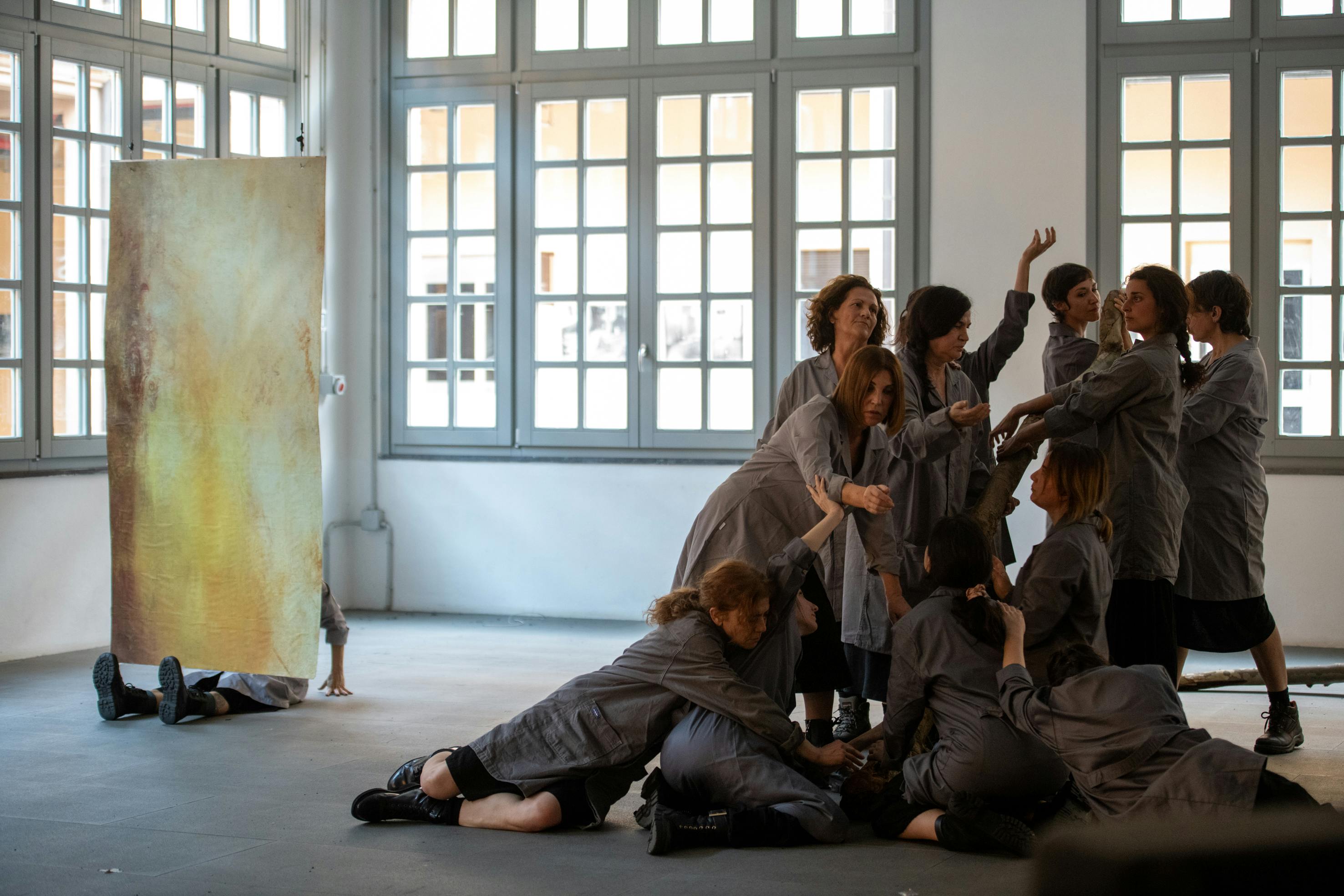 A group of women in gray coats and combat boots are gathered to form almost a tableau vivant. The performers are in a bright industrial space with large windows