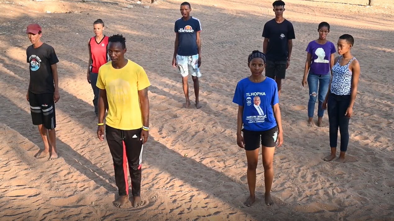 A group of eight performers standing with their legs together and their arms at their sides are on sandy ground