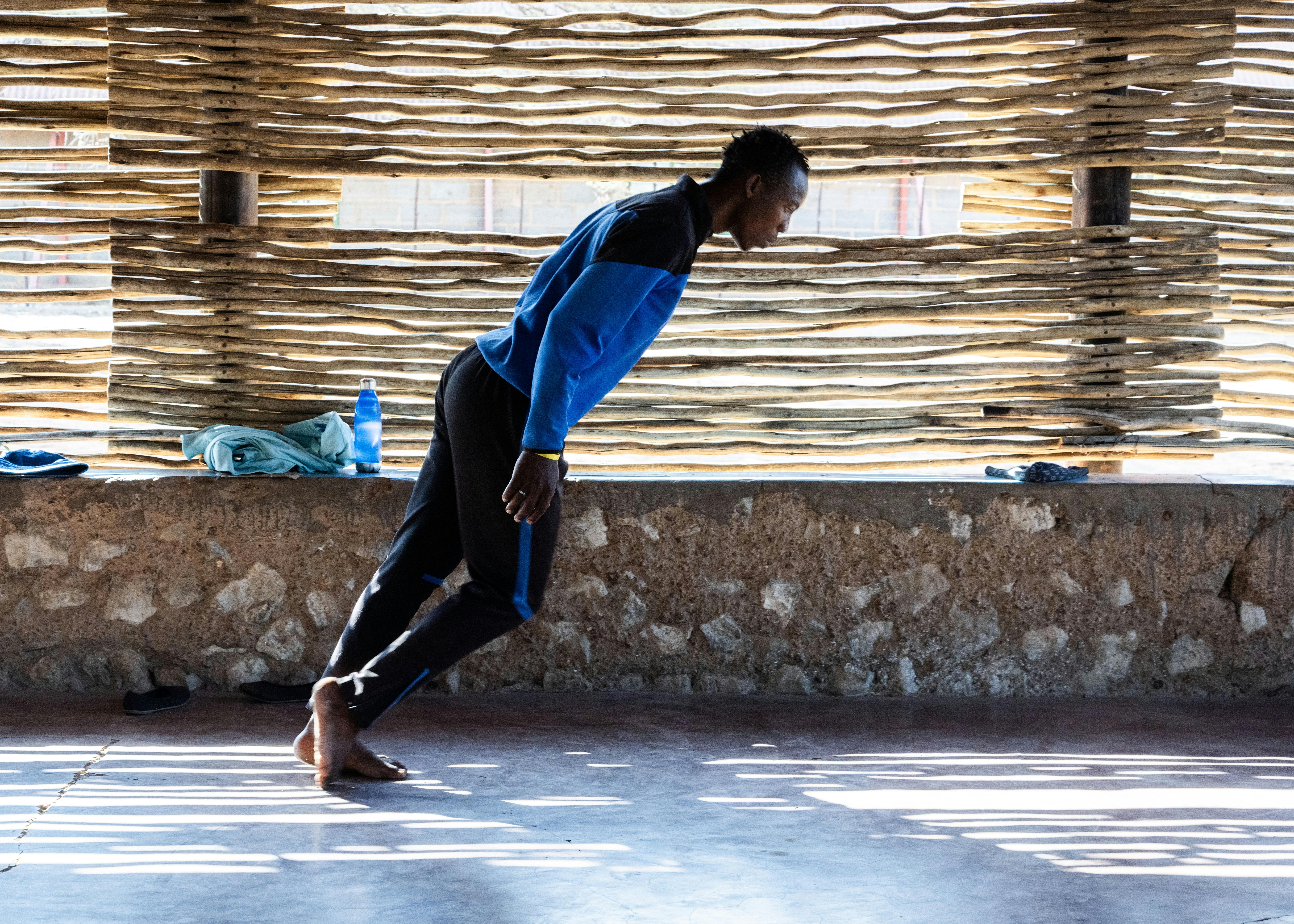 A performer in a blue and black suit has his body straight and leaning forward, his arms at his sides. He is in a room with a low stone wall and bamboo canes to shield the light
