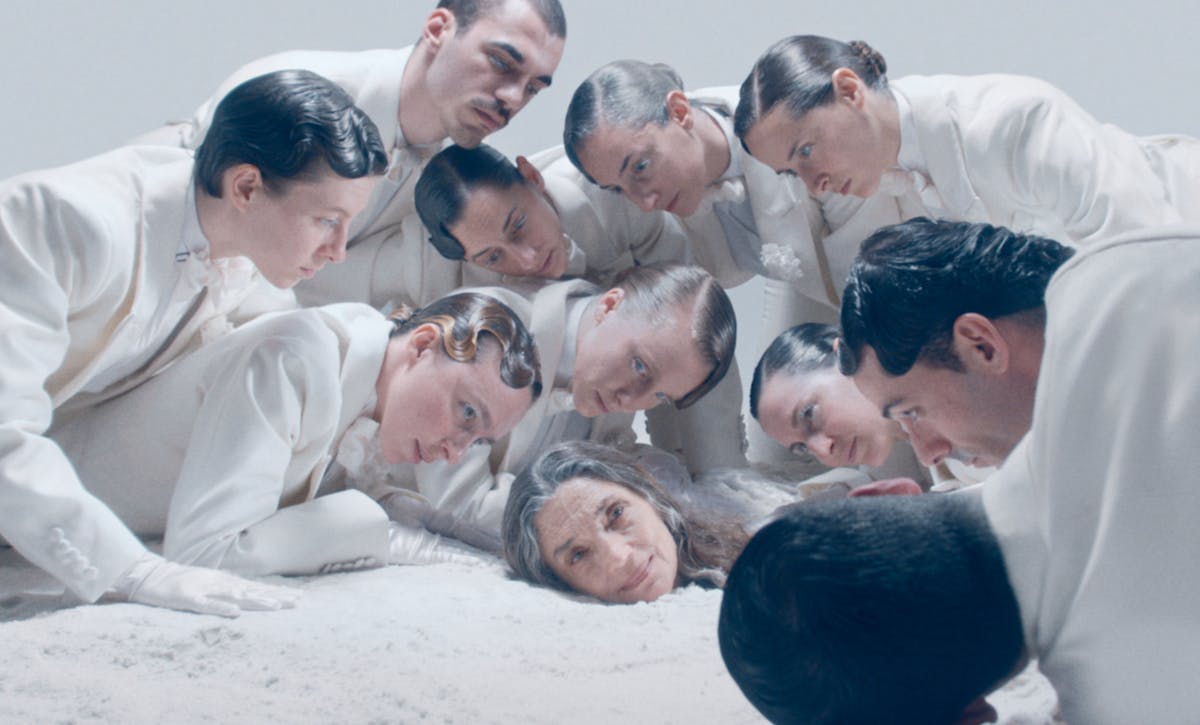 Undici performer sono sdraiati a terra, uno sopra l'altro. Sono all'interno di uno spazio bianco e anch'essi vestiti di bianco, con i capelli raccolti e cosparsi di gel. Dieci di loro, più giovani, osservano la più anziana che è sdraiata sotto a tutti, di cui si vede solo il viso