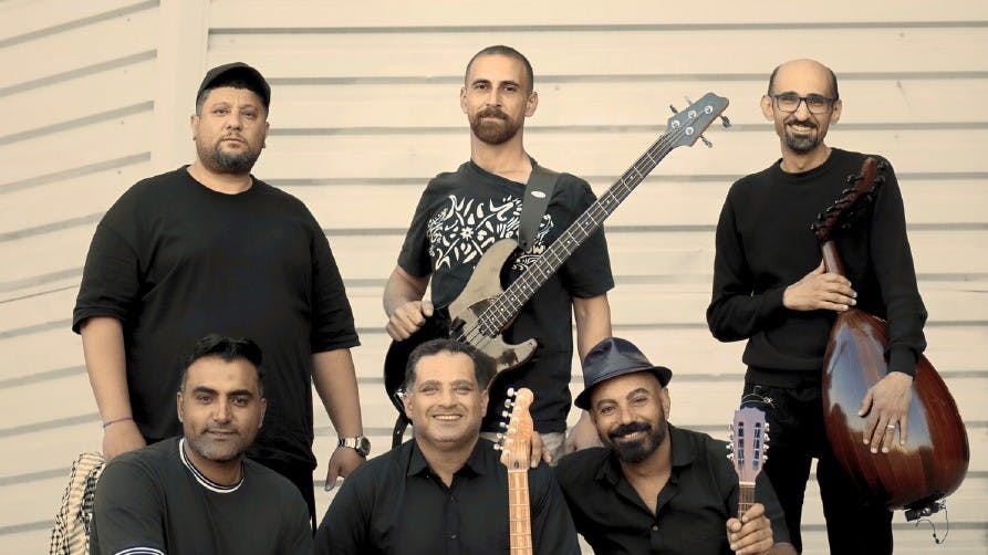La band Radio Gaza Six men, three standing, three kneeling, smile with their musical instruments in their hands: percussions, guitars, traditional instruments of the Middle East.