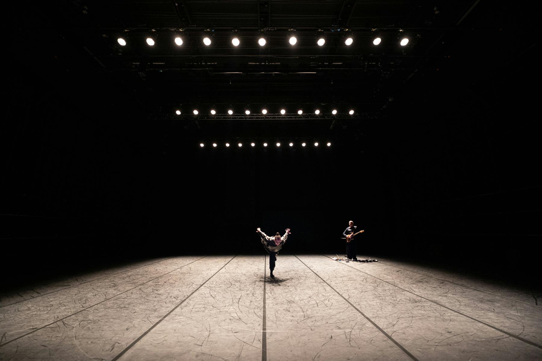 In a dark and bare theatrical space, illuminated from above by three rows of spotlights, two figures. A female, in the center, stands on one leg, with her body bent forward and her arms stretched backwards. The other, in the background on the right, has a guitar in her arms.