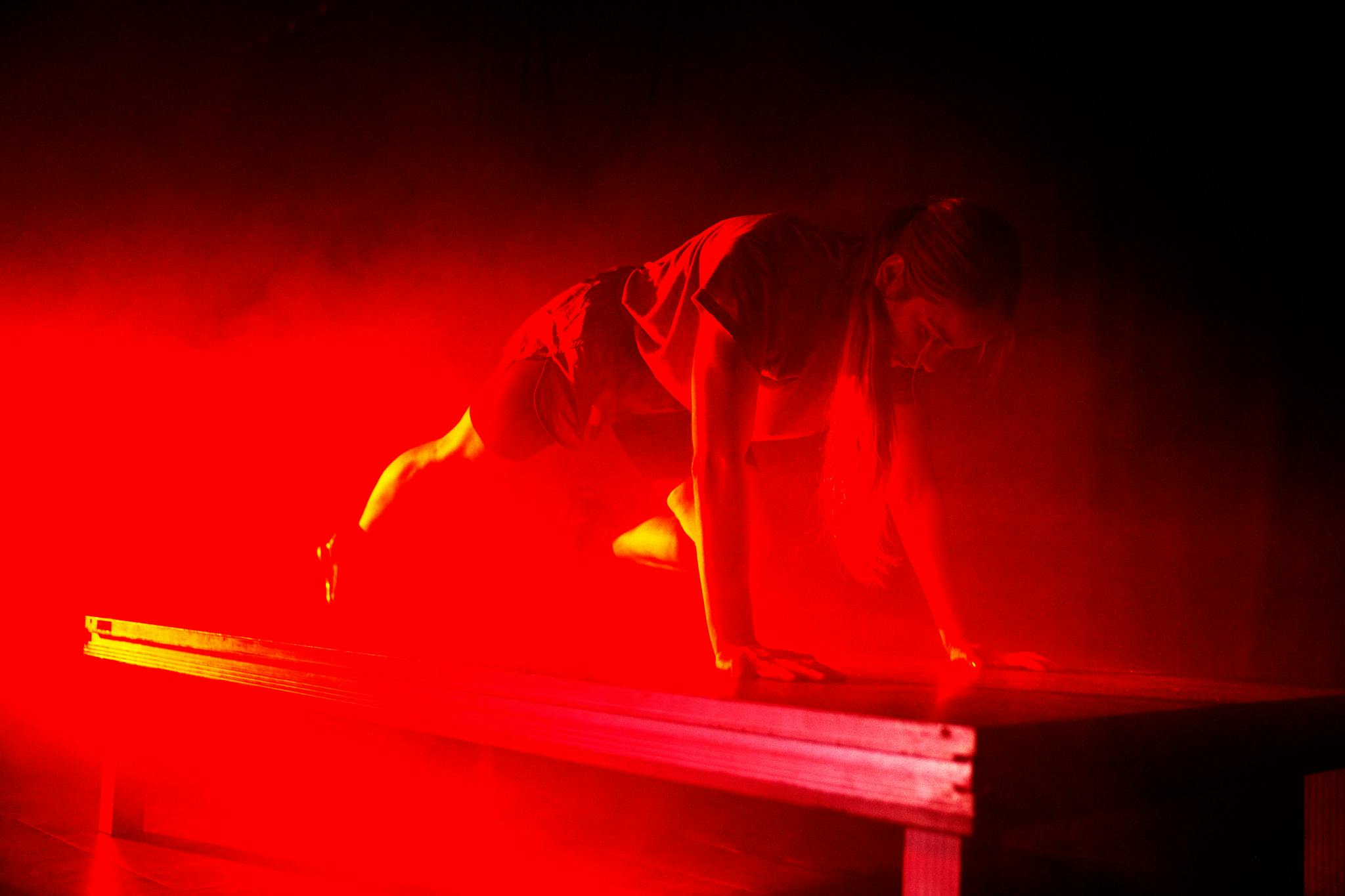Fotografia con luce molto rossa di una danzatrice col corpo allineato, dal capo ai piedi, in posizione orizzontale, poggiando su avambracci e punte dei piedi