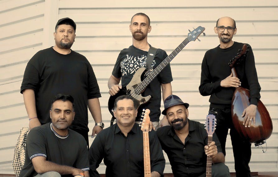 Six men, three standing, three kneeling, smile with their musical instruments in their hands: percussions, guitars, traditional instruments of the Middle East.