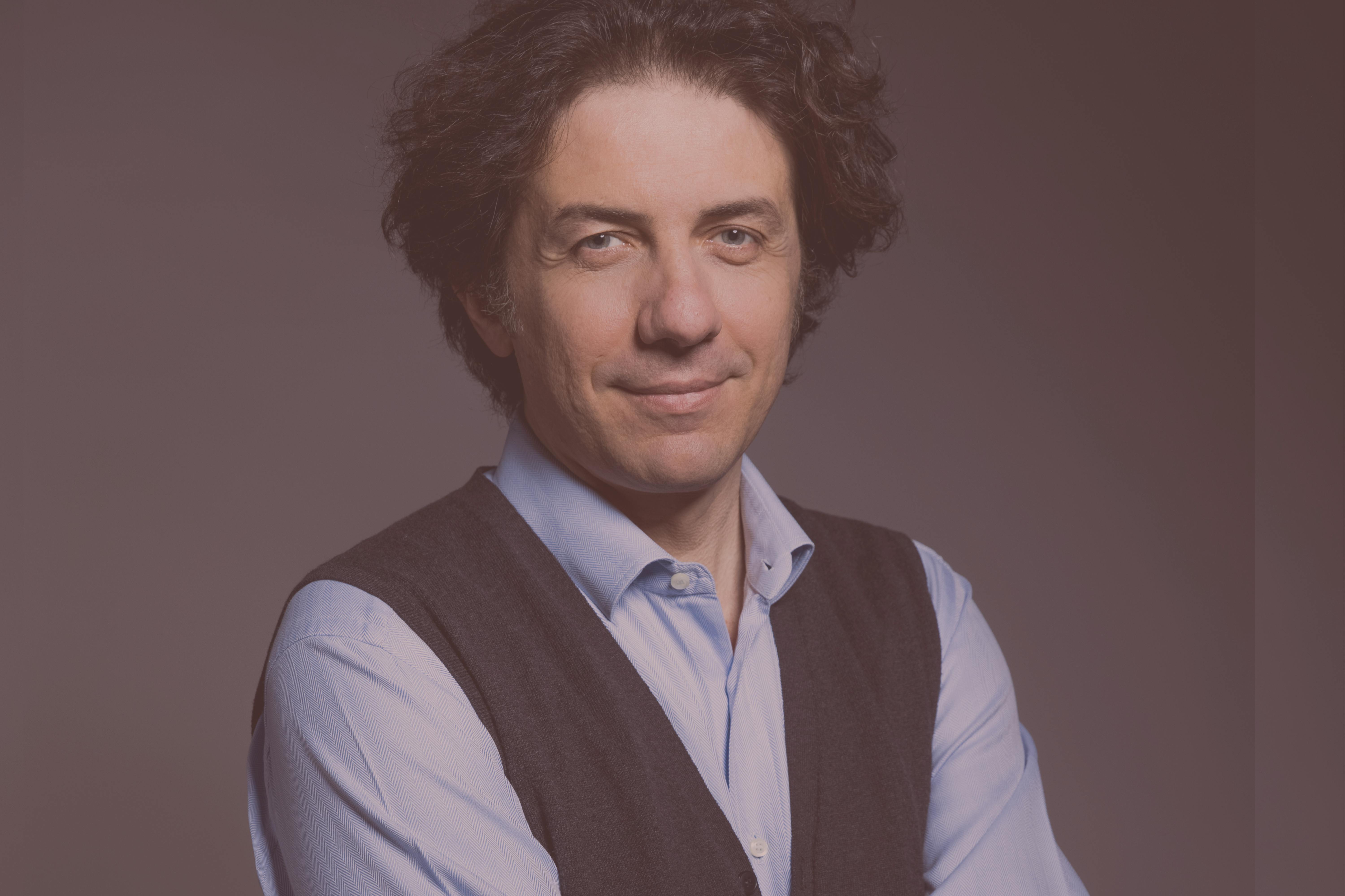 Marco Cappato con i capelli ricci, indossa una camicia sui toni dell'azzurro e un gilet scuro