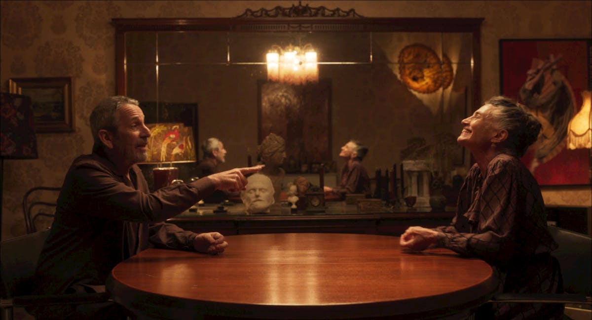 In una sala da pranzo due persone sono sedute ai lati opposti del tavolo, la figura sulla sinistra indica con un dito quella a destra, che sorride.