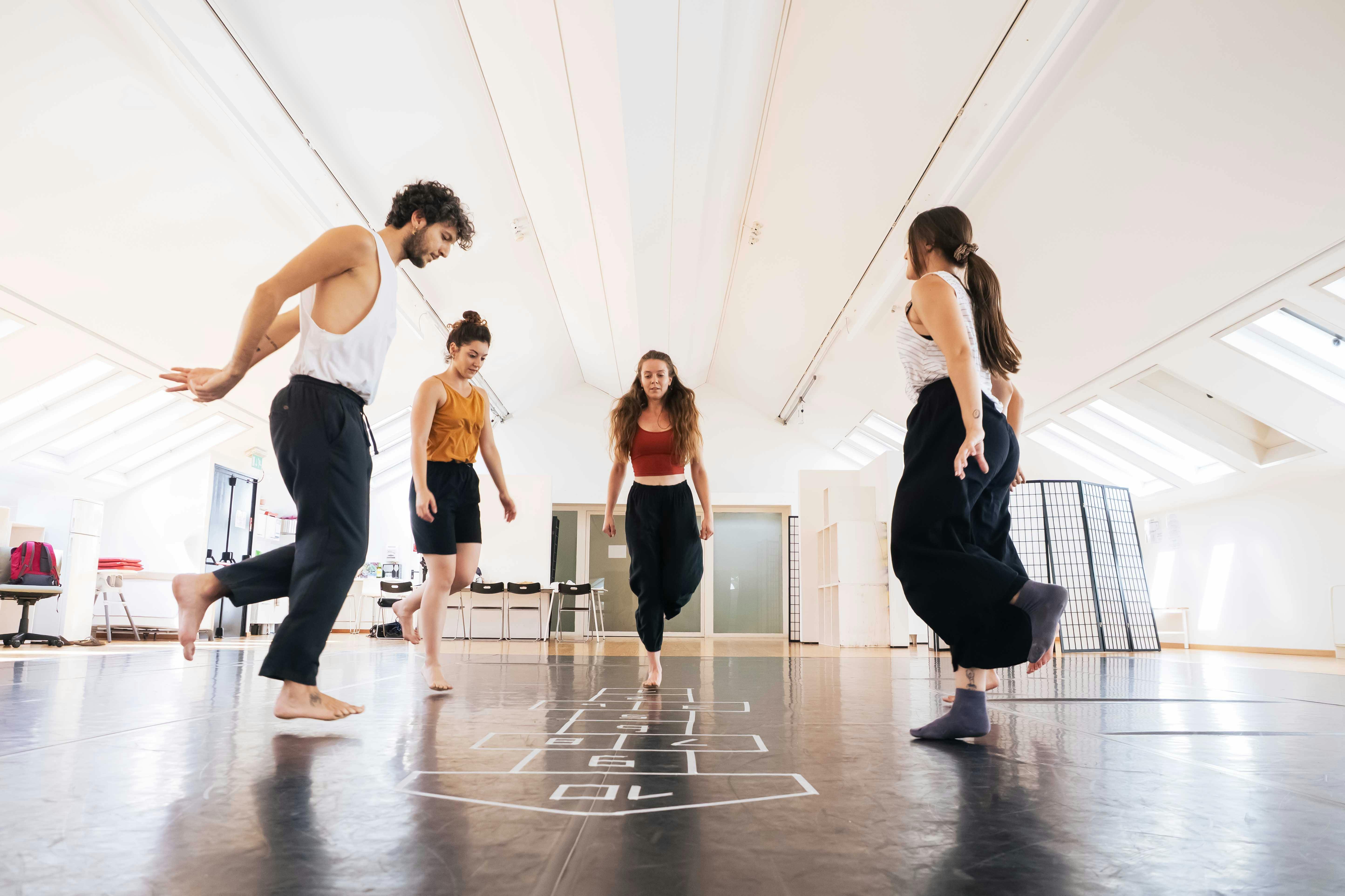 Sul tappeto danza è disegnato in bianco il gioco della campana. Intorno saltano su un piede solo cinque performer, una delle quali è nascosta e se ne intravede solo il piede. L'ambiente è uno studio danza