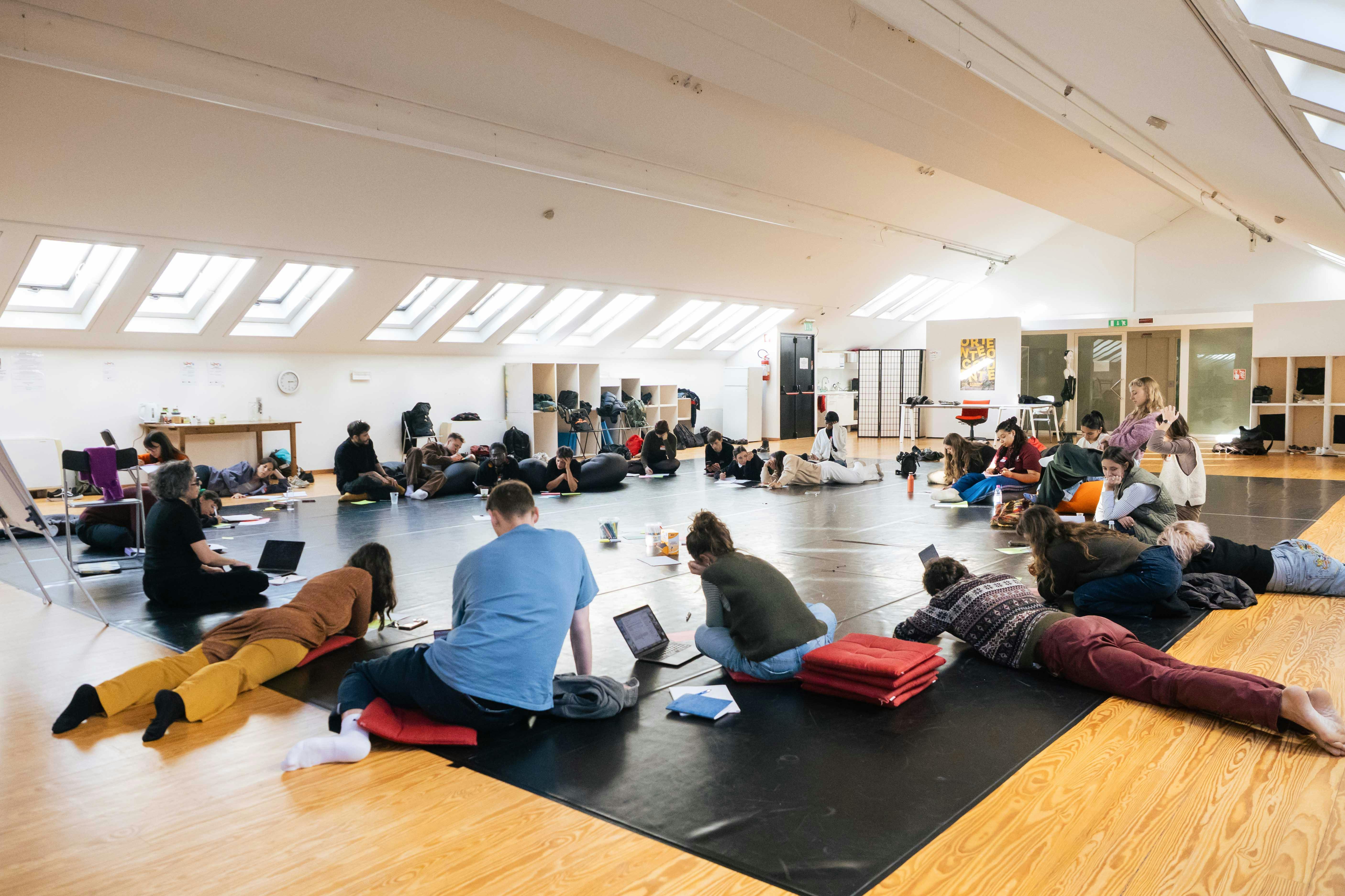 Partecipanti all'Atelier al lavoro in sala, il gruppo è in cerchio alcune persone sono sedute per terra, altre sdraiate