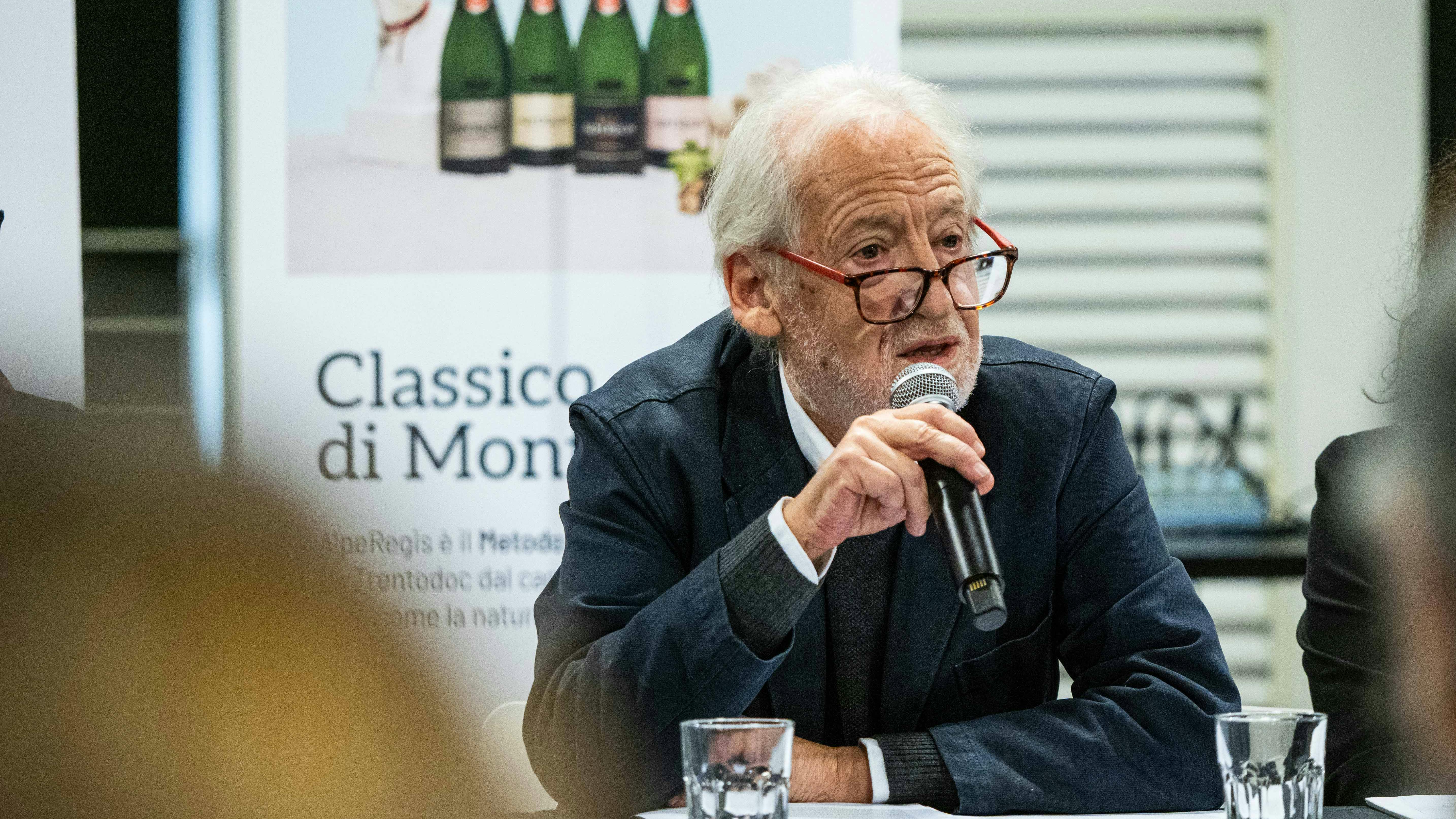 Lanfranco Cis, co-fondatore di Oriente Occidente e direttore artistico dal 1981 | ph. Giulia Lenzi Lanfranco Cis è un uomo bianco sulla settantina, con i capelli bianchi e la barba bianca. Indossa una giacca scura e occhiali con le stanghette rosse. Ha in mano un microfono e sta parlando durante un evento