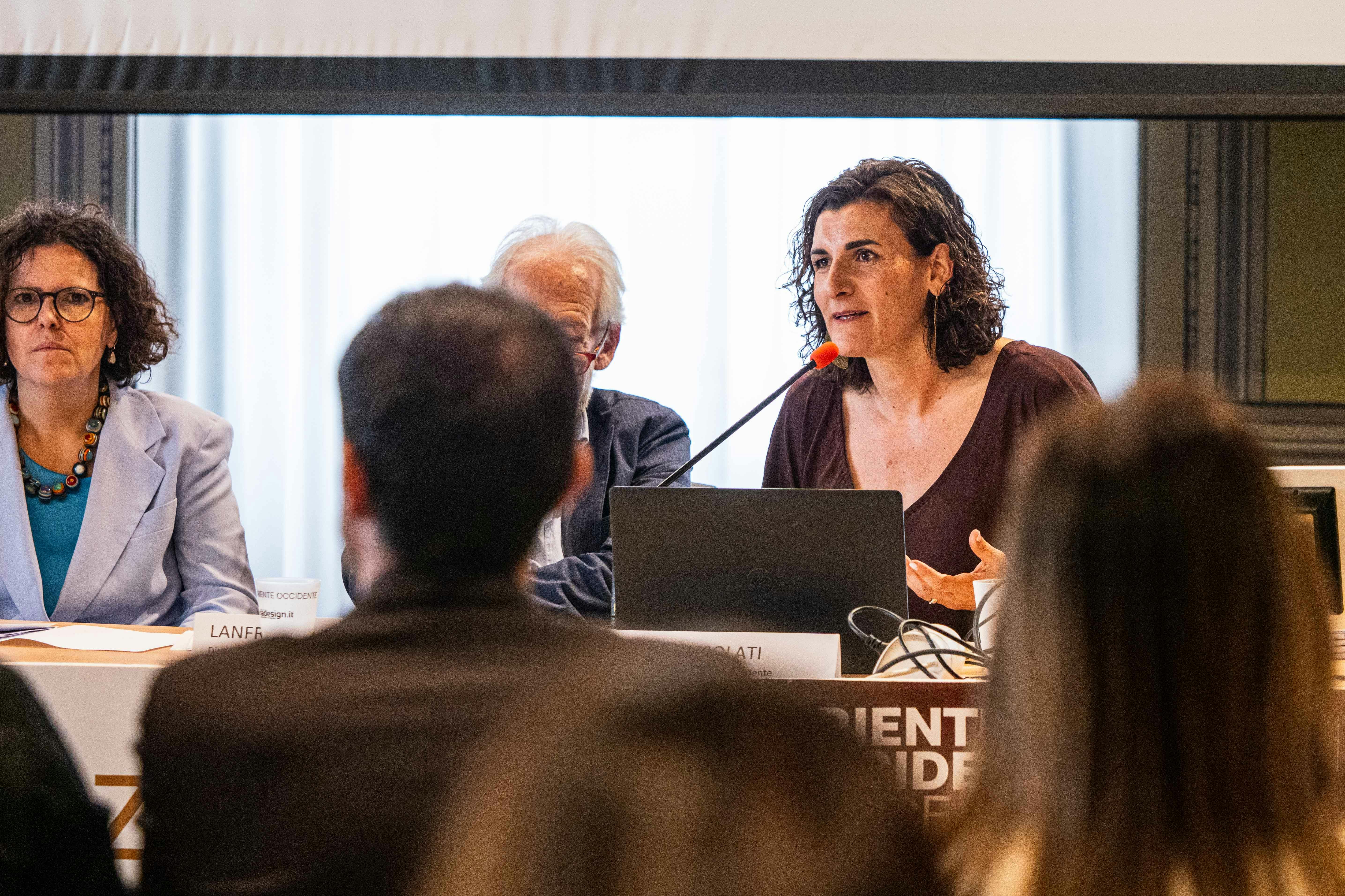 Anna Consolati è una donna bianca, con capelli ricci lunghi fino alle spalle. In questa foto indossa un vestito marron e parla al microfono seduta a un tavolo con un computer davanti a sé