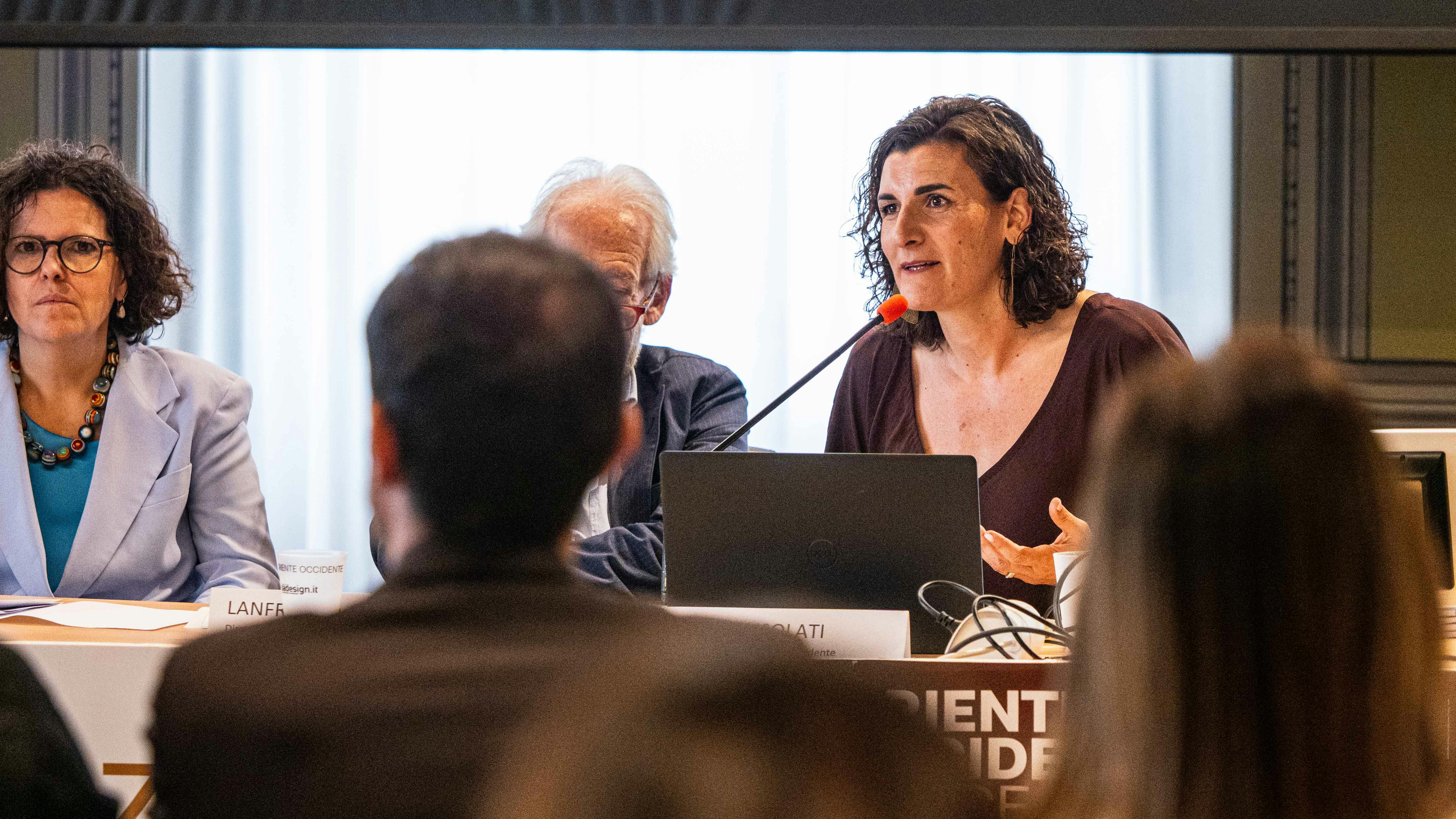 Anna Consolati, direttrice generale di Oriente Occidente Anna Consolati è una donna bianca, con capelli ricci lunghi fino alle spalle. In questa foto indossa un vestito marron e parla al microfono seduta a un tavolo con un computer davanti a sé