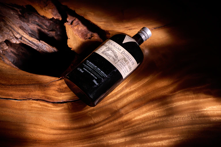 A rare bottle of The Macallan 1801 Replica rests on a polished wooden surface, warmly lit to highlight its vintage charm. This collectible whisky embodies craftsmanship and legacy.