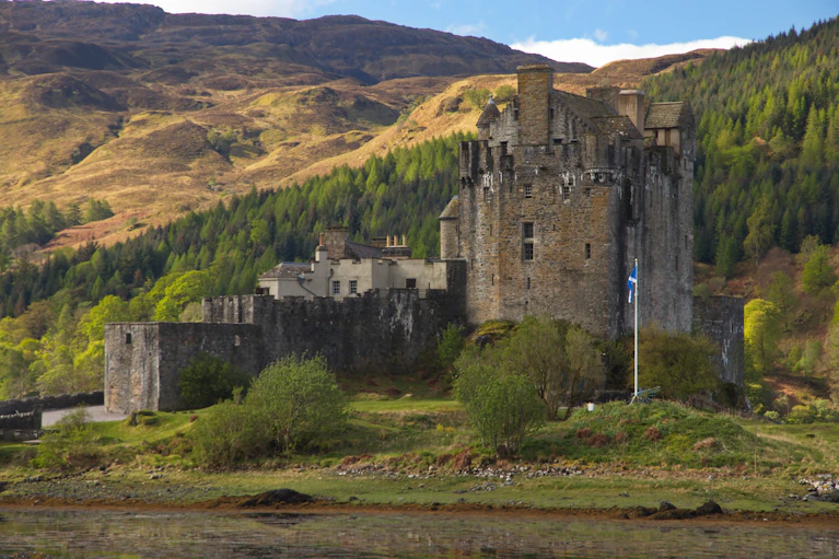 A medieval castle set in the Scottish Highlands, surrounded by green hills and with the Scottish flag in the foreground. The scene evokes the deep cultural roots that underpin the long-term value of Scotch single malt whisky as an investment.