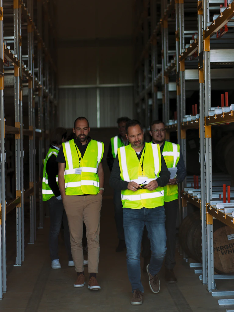 A group of investors wearing high-visibility safety vests walking through the Scotch Whisky Investments bonded warehouse in Glenrothes, Scotland, viewing racks filled with maturing whisky casks.