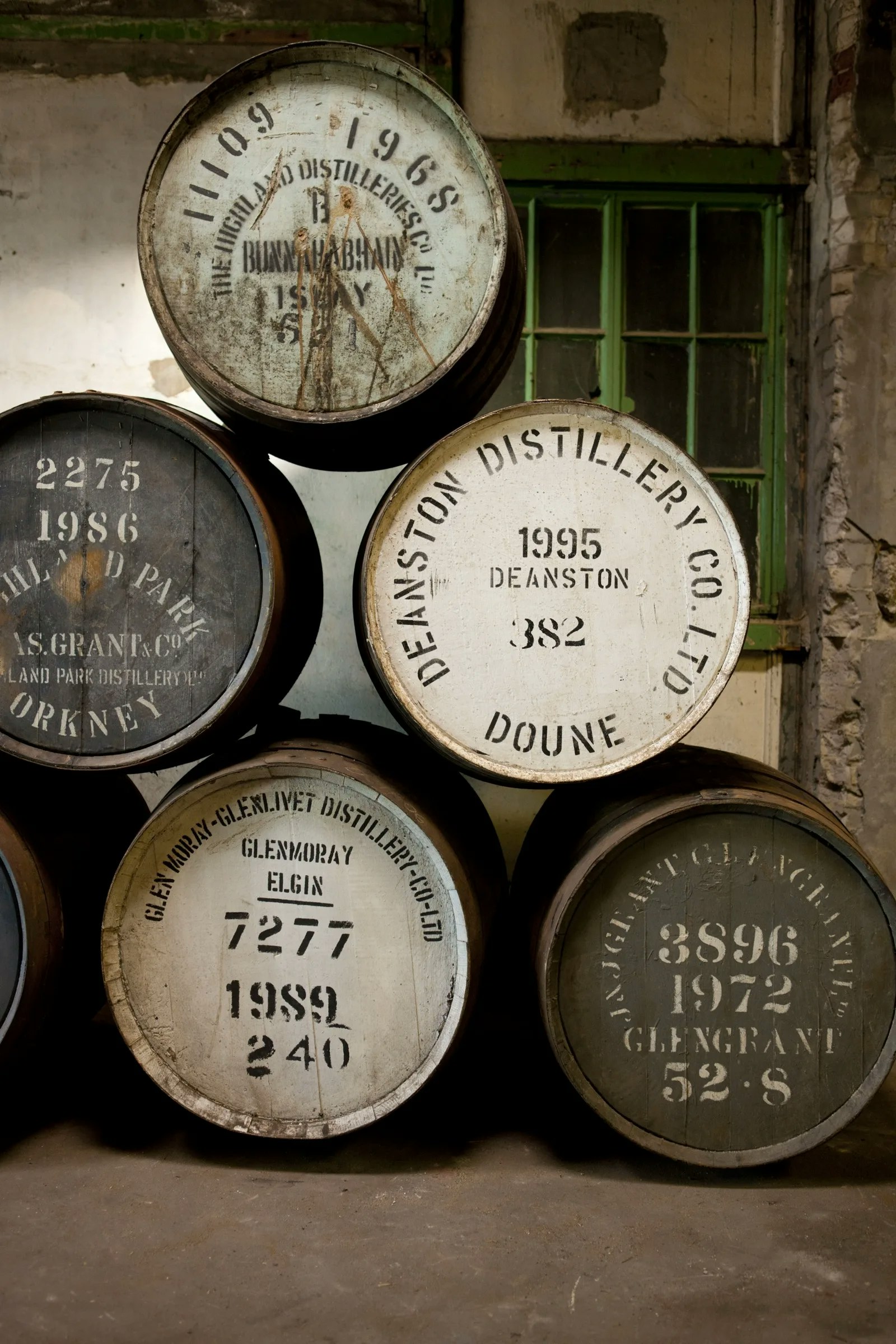 Gestapelde whiskyvaten met distilleerderijmerken in een verweerd warehouse