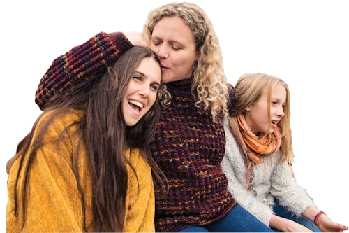 A woman sits in between two young girls. She leans into one of the girls and kisses her forehead