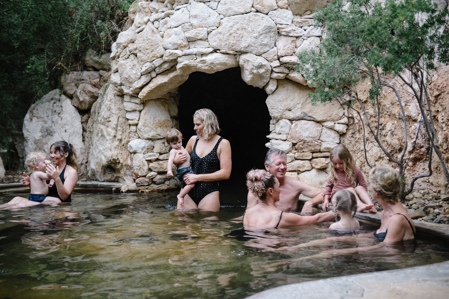 group of women and children bathing in geothermal baths with the opening of a cave in the background