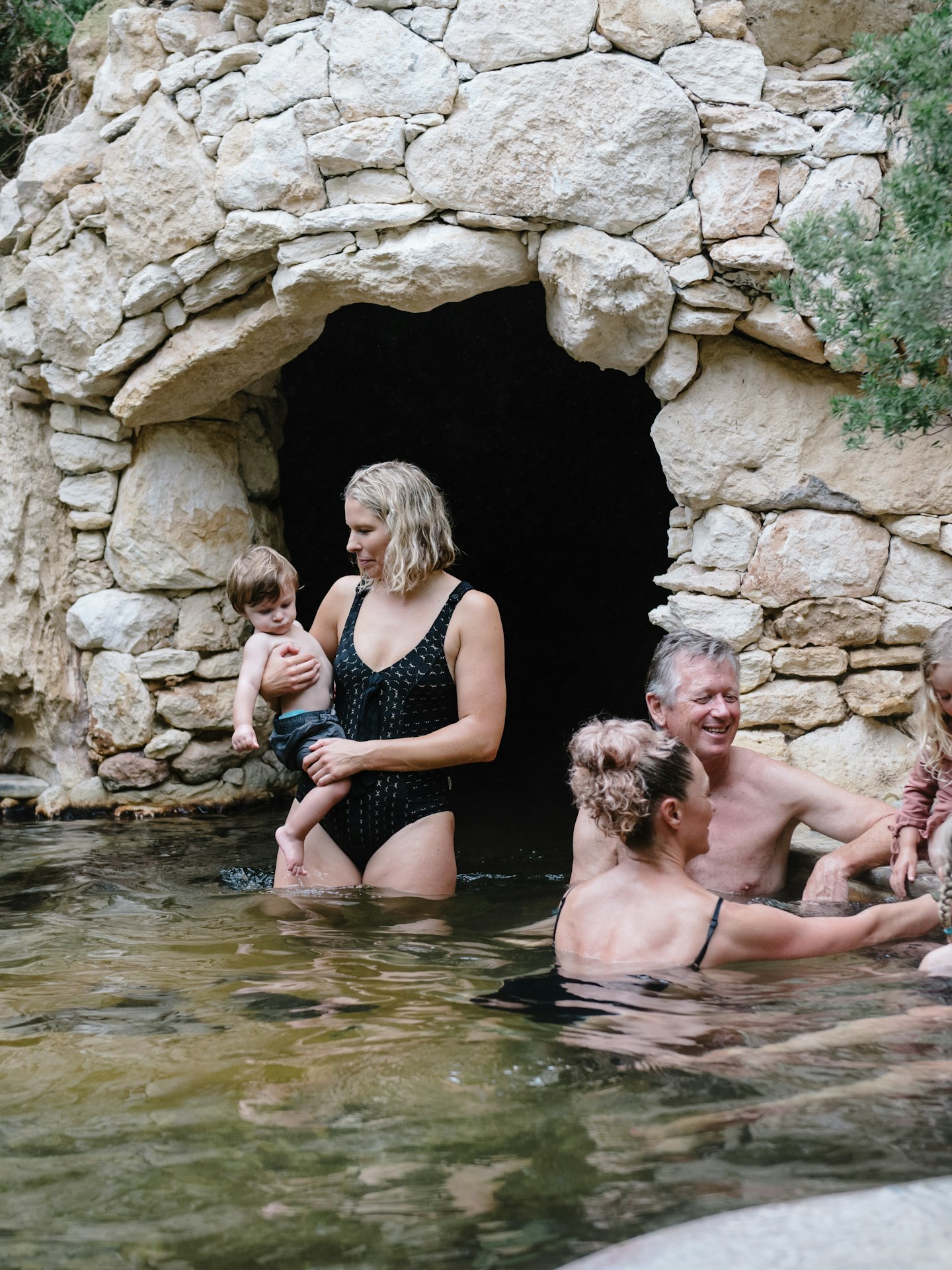 group of women and children bathing in geothermal baths with the opening of a cave in the background