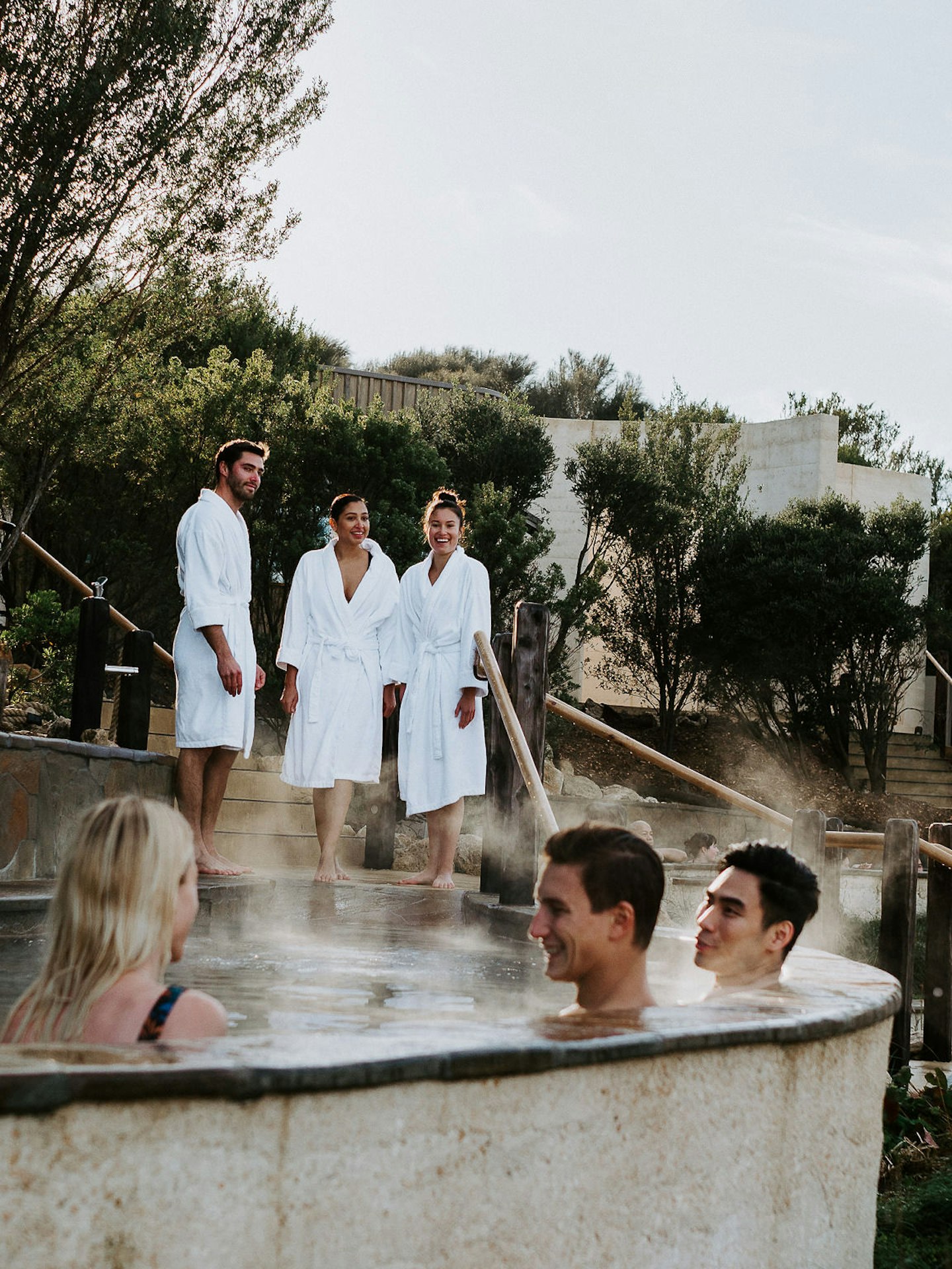 three friends sitting in Amphitheatre hot pool and three friends in white bath robes standing behind