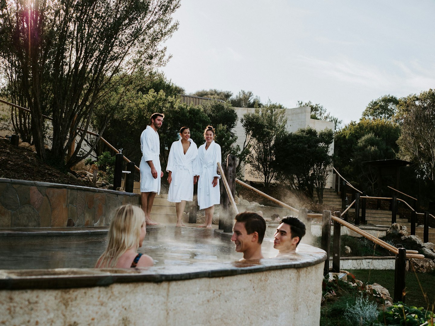 three friends sitting in Amphitheatre hot pool and three friends in white bath robes standing behind