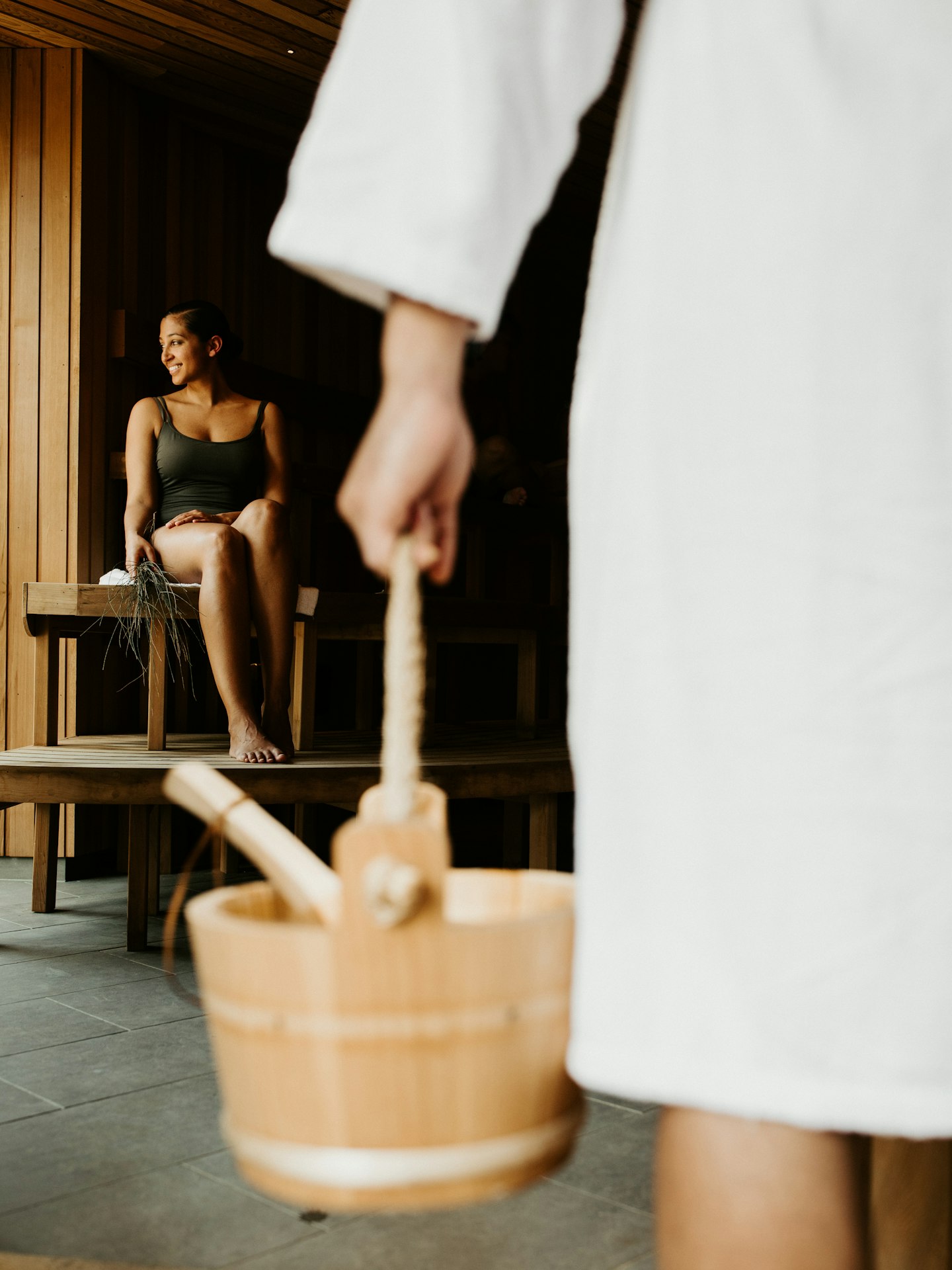 woman sitting in sauna and man in white bath robe in foreground carrying wooden bucket