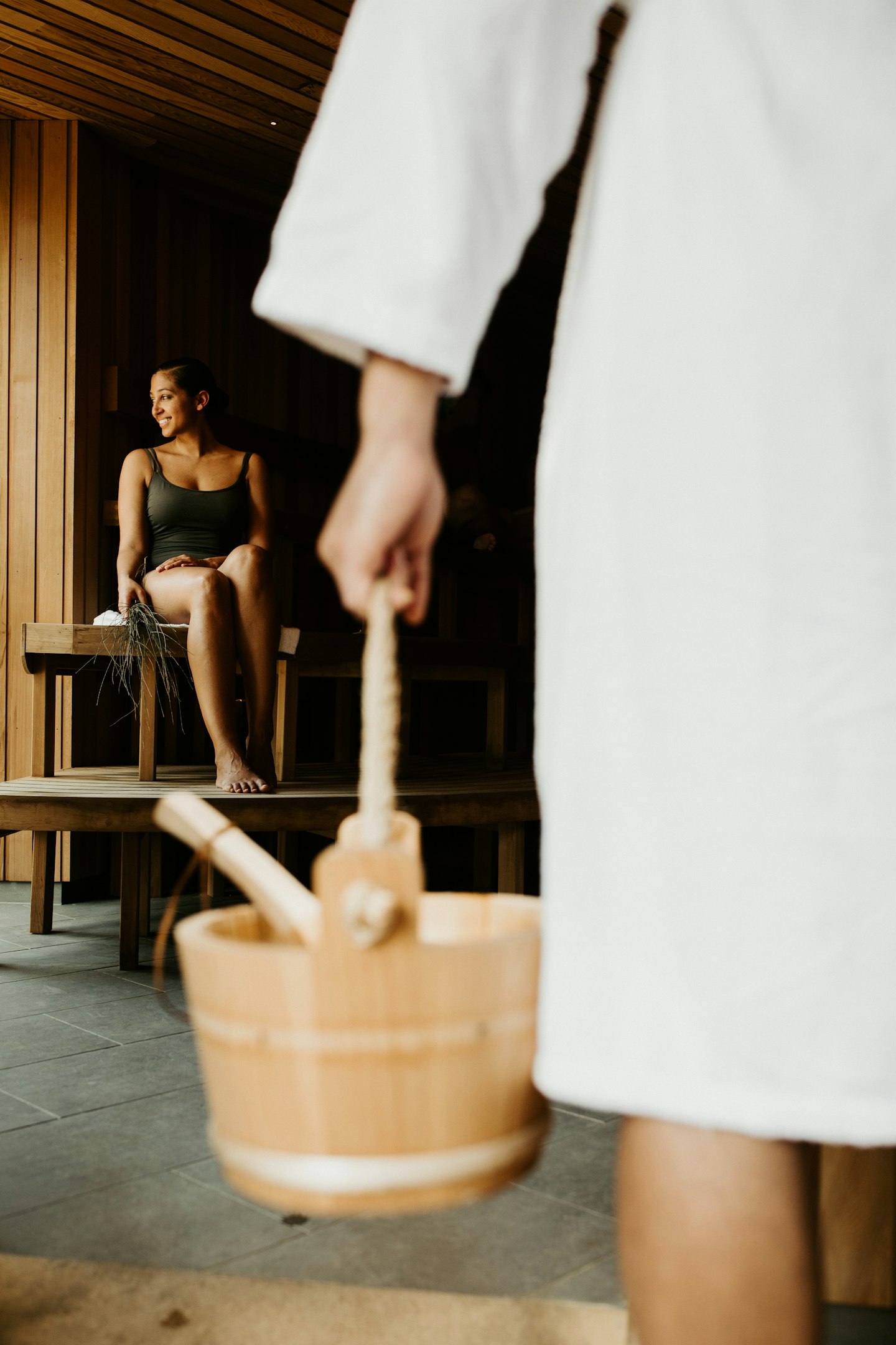 woman sitting in sauna and man in white bath robe in foreground carrying wooden bucket