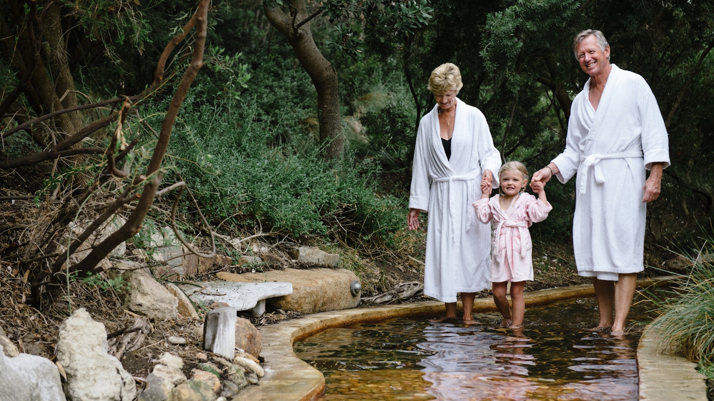 grandparents in white bath robes holding hands with granddaughter in pink bath robe, standing on reflexology walk.