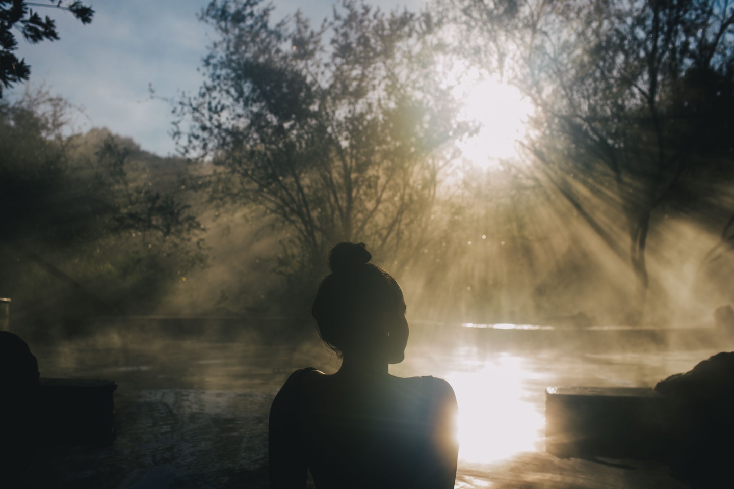 woman sitting in steamy hot pool with sun breaking through the trees