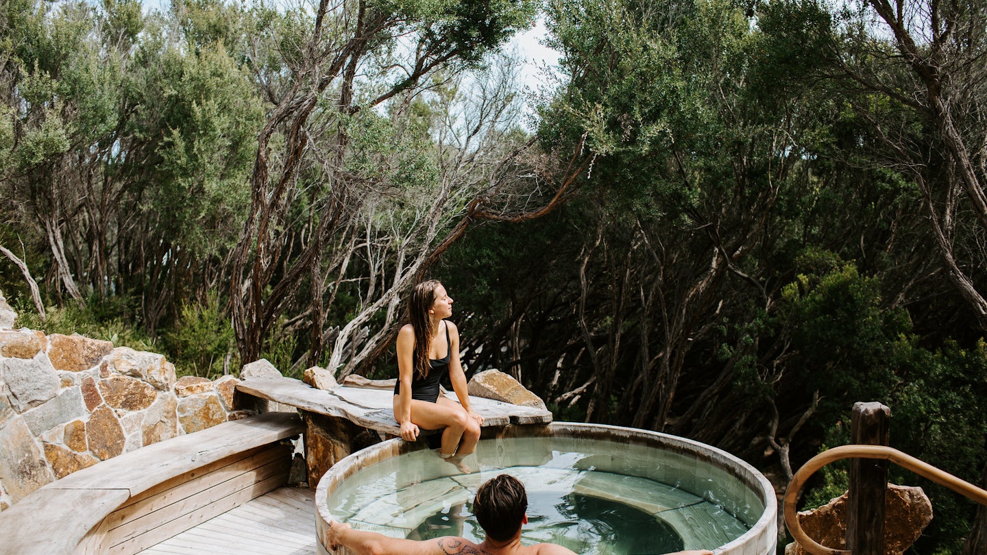 woman sitting on the ledge of barrel pool with a man soaking in it