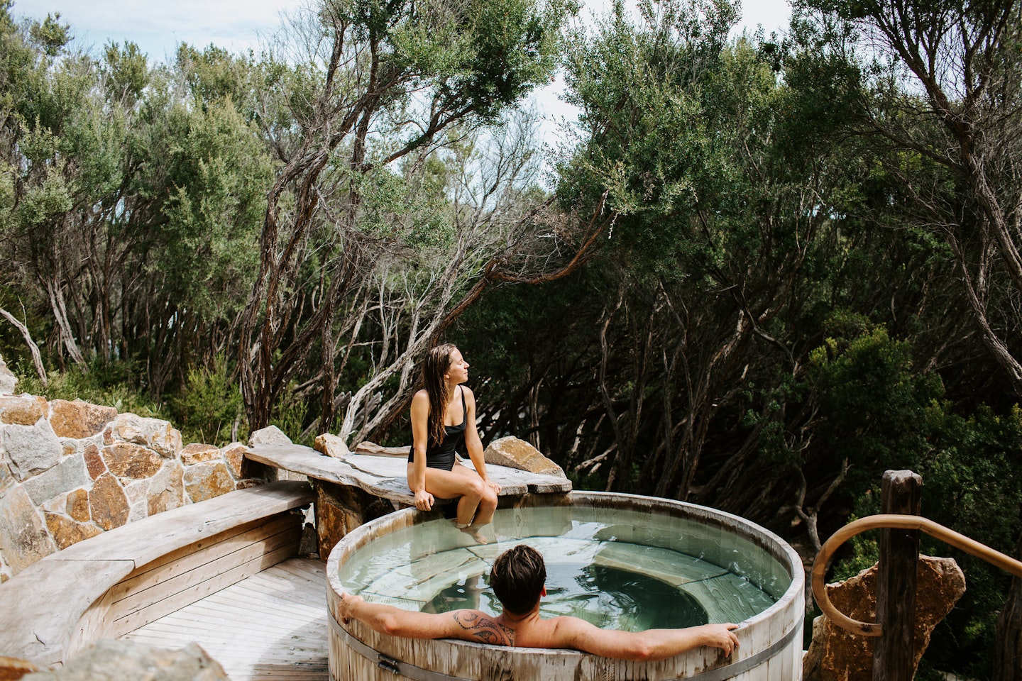woman sitting on the ledge of barrel pool with a man soaking in it