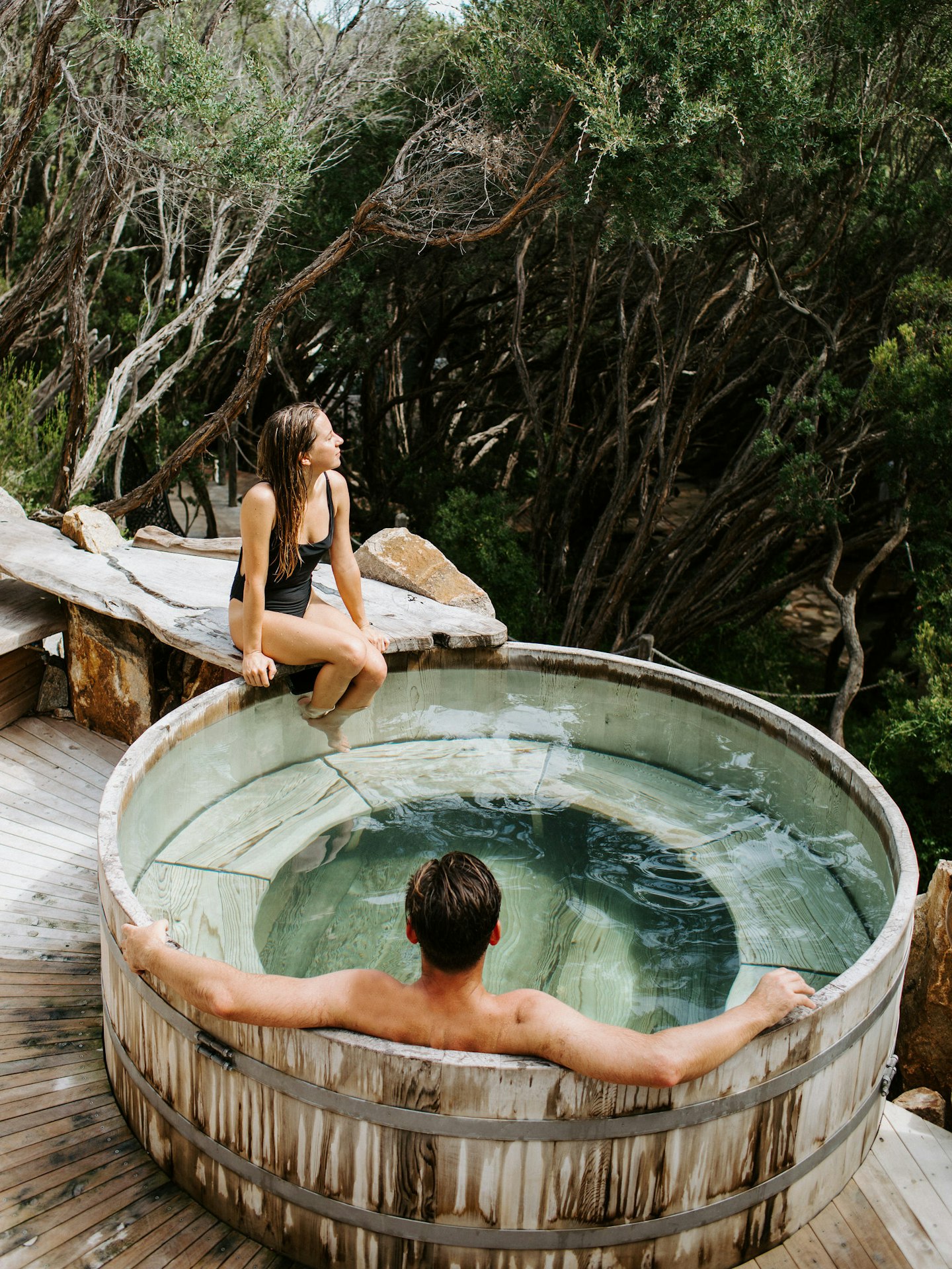 man sitting in barrel pool and woman sitting on edge looking out at trees