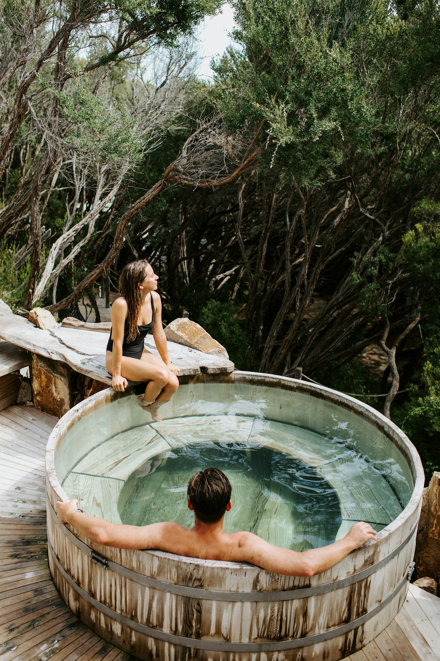 man sitting in barrel pool and woman sitting on edge looking out at trees